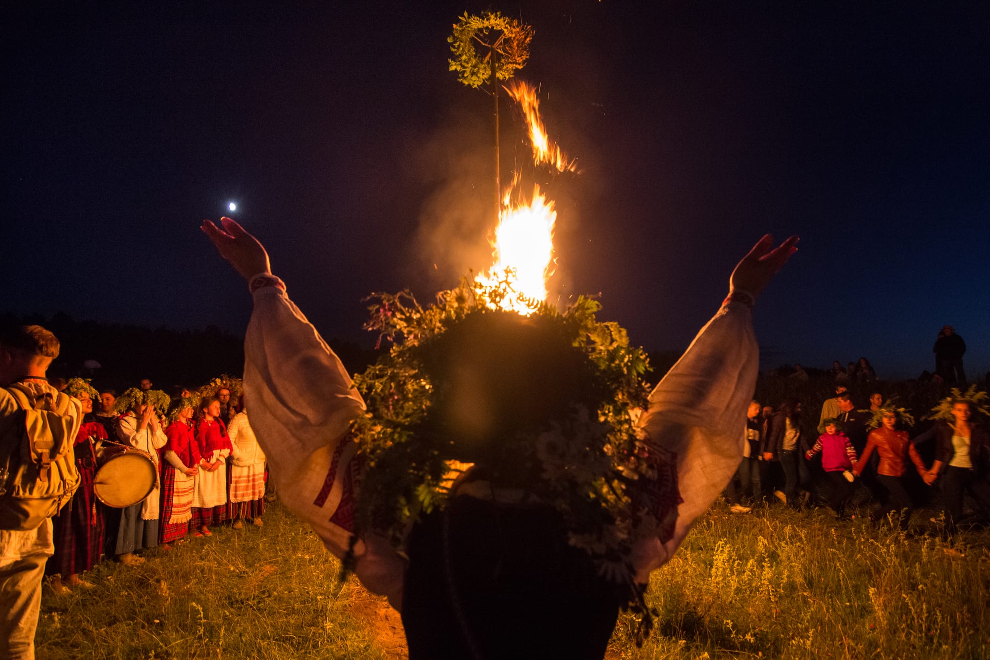 Seen from behind, a woman in an embroidered white shirt and a large wreath of greenery raises both arms towards a tall bonfire at night, with people in folk dress and a drummer gathered to the left