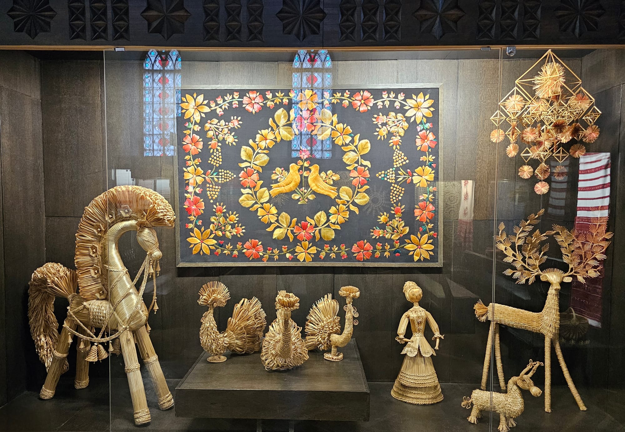 A museum display case of golden straw figures, including a large horse, birds, a deer and a woman, in front of a dark panel decorated with straw flowers and two birds, with a hanging geometric straw ornament to the right