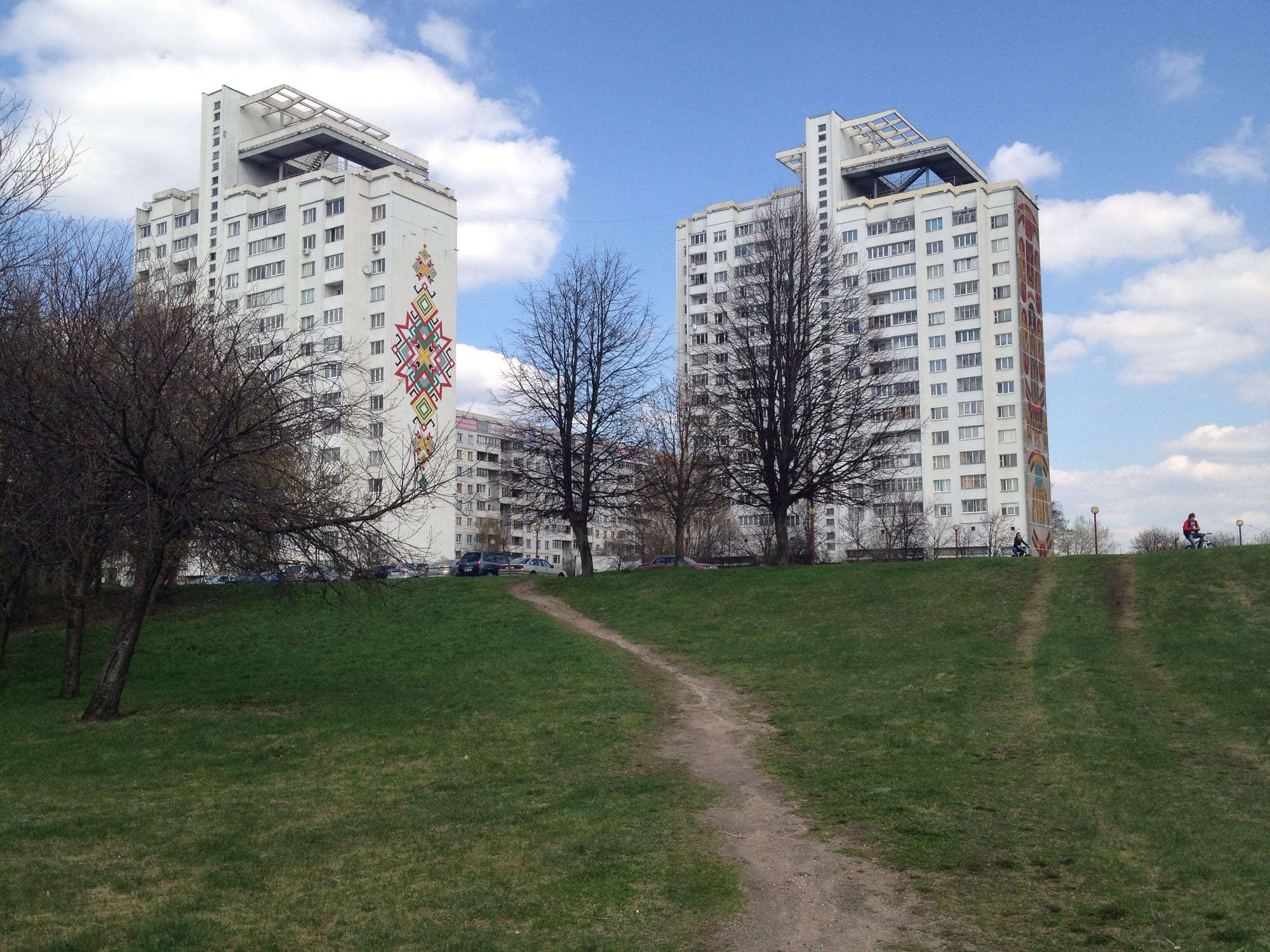 Two tall Minsk apartment blocks decorated with a traditional woven-pattern mural, seen across a grassy park with a footpath and a cyclist