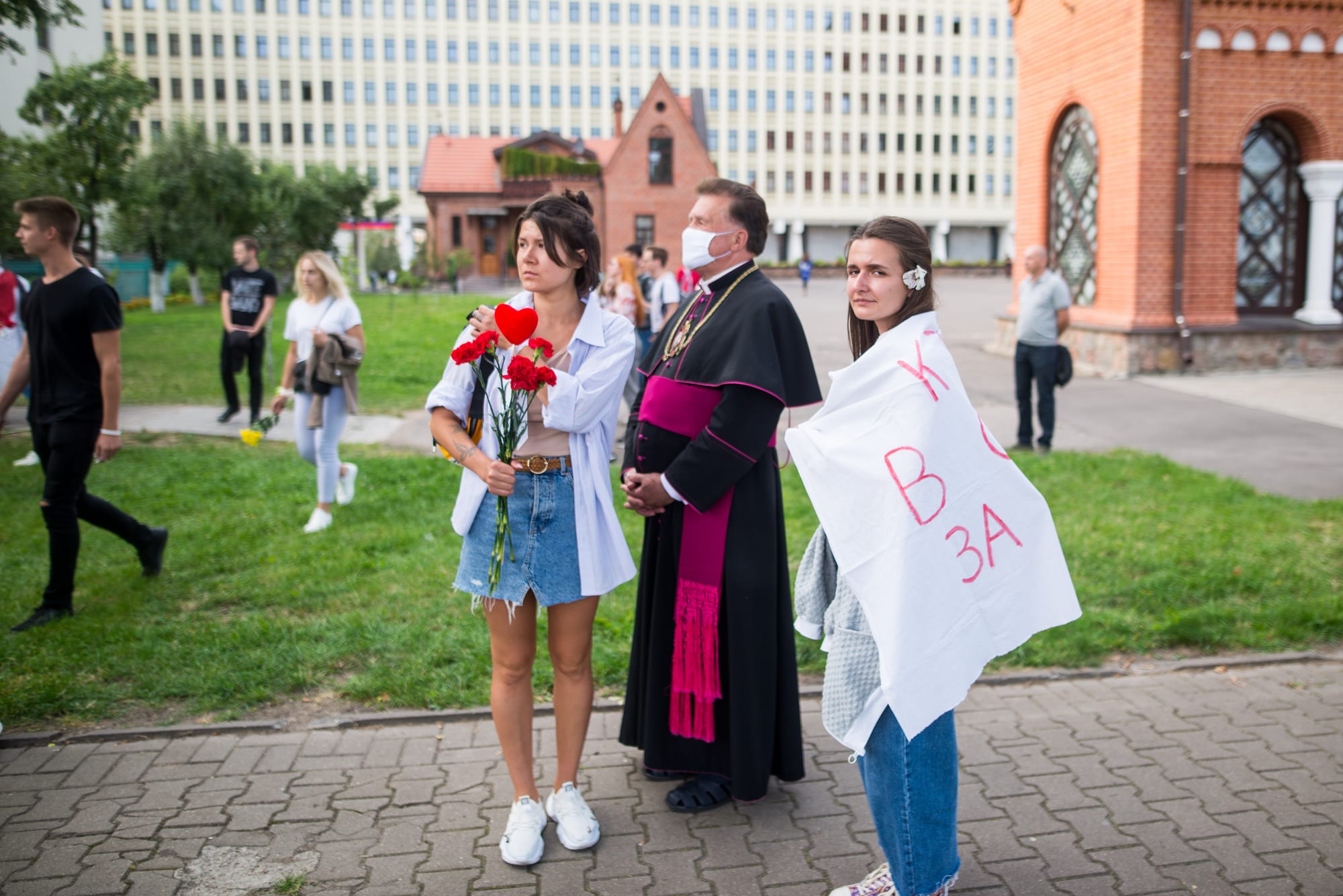 A vast crowd carrying white-red-white flags filling a Minsk avenue past a red-brick church