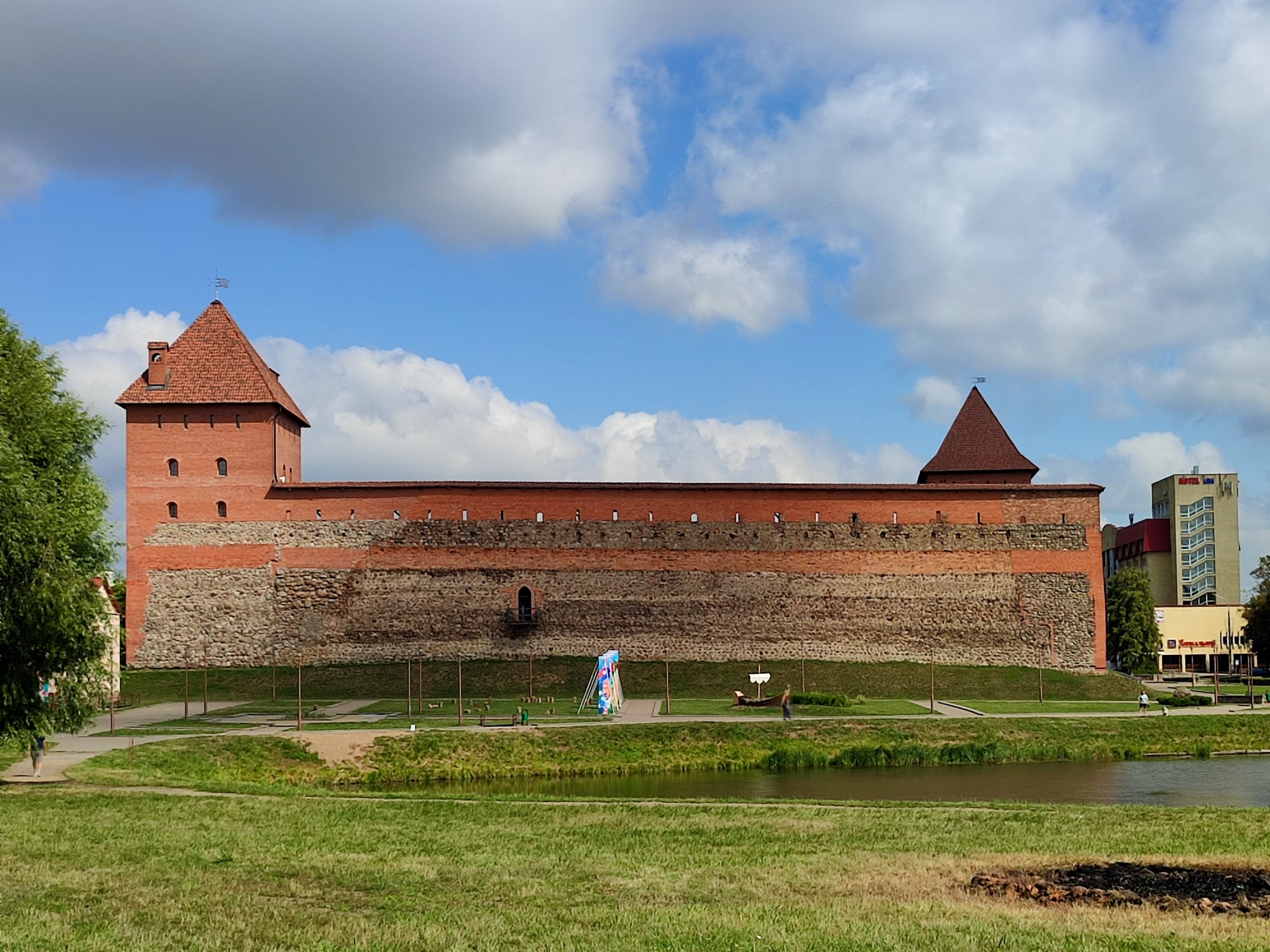 A long fortress wall of fieldstone topped with red brick, with two square red-roofed towers, seen across a pond and grass under a cloudy blue sky