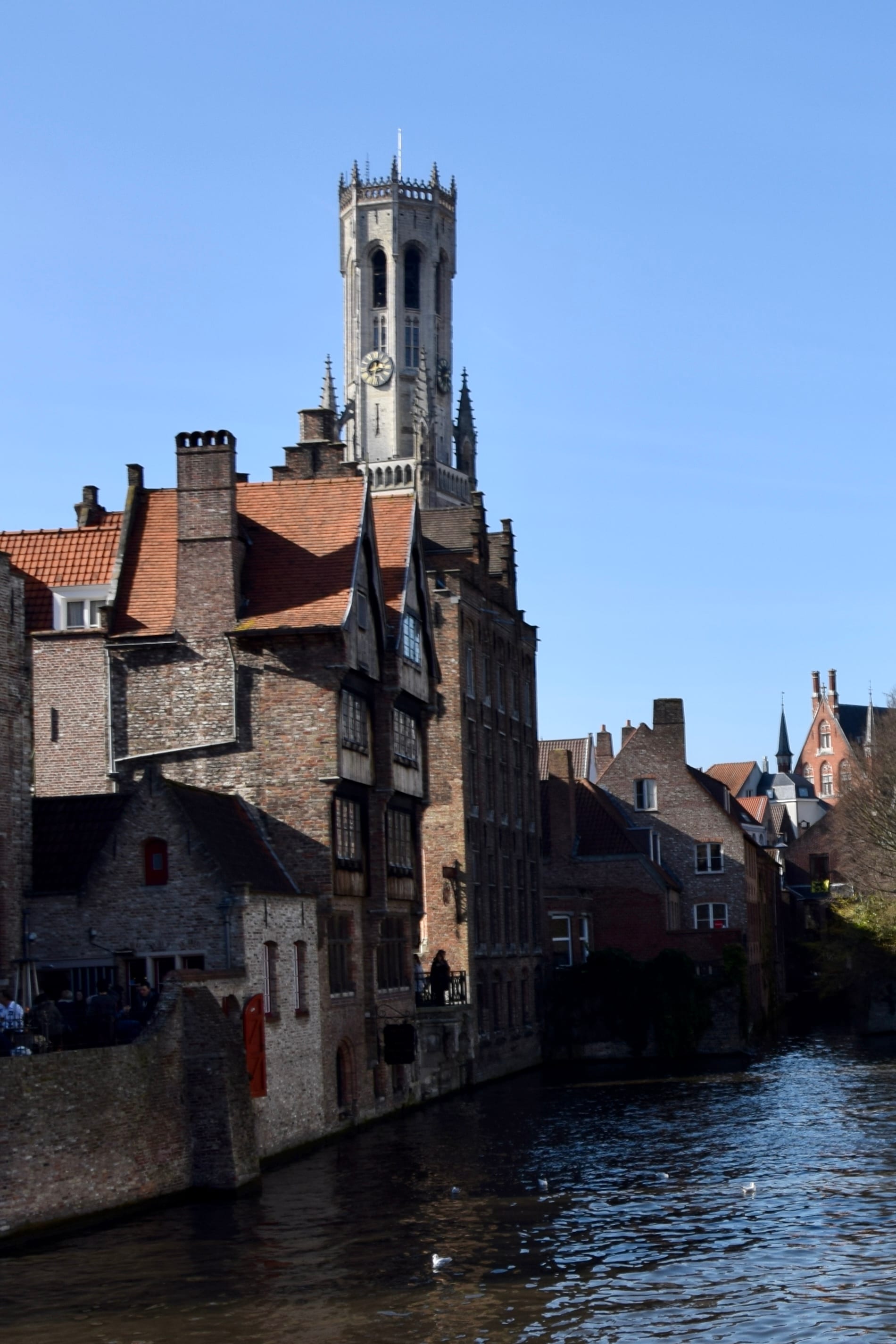 A stone canal quay with stepped-gable houses and a tall medieval belfry behind, reflected in still water