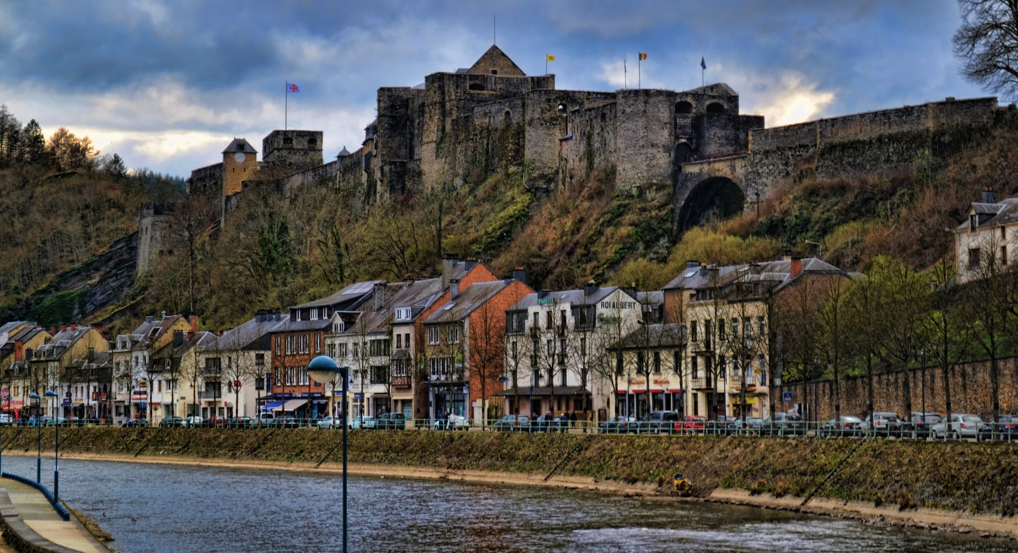 A long stone castle on a rocky ridge above a town wrapped in a river bend, forested hills around