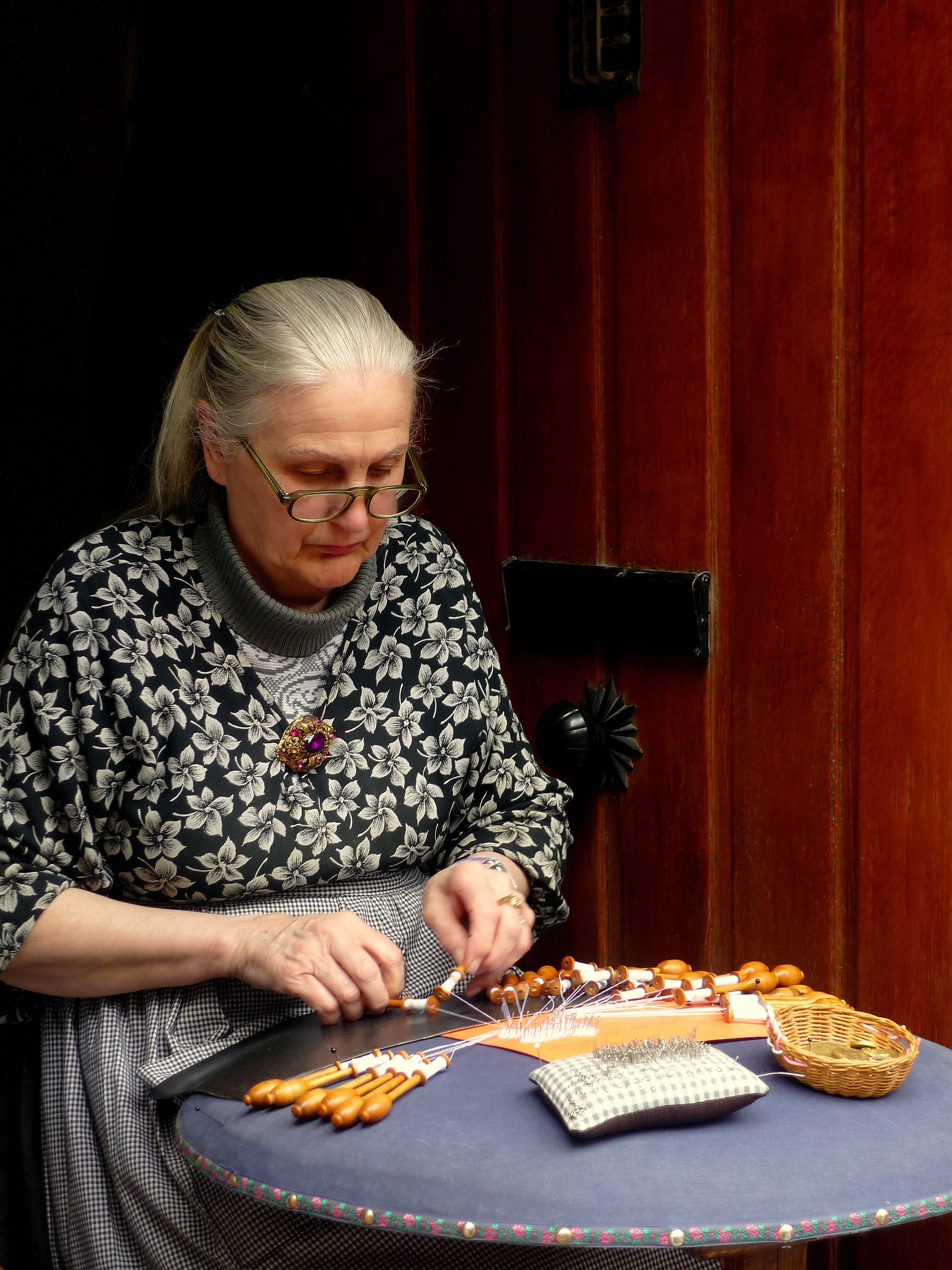 A grey-haired woman in glasses and a patterned dress works bobbin lace at a small round table, with many wooden bobbins and a pincushion spread in front of her