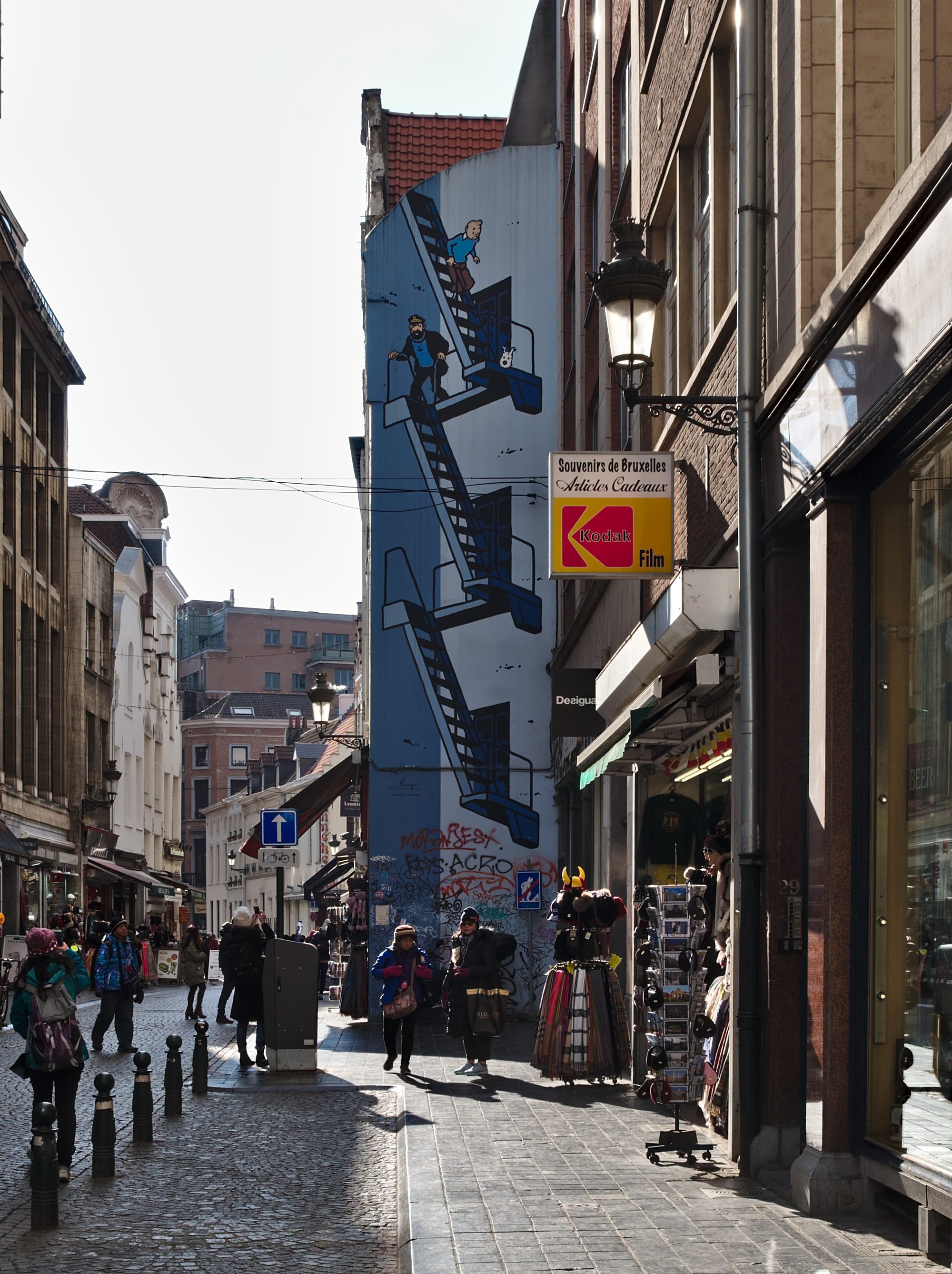 A tall blue gable wall painted with a comic-book scene of Tintin, Captain Haddock and Snowy on a zigzagging fire escape, above a cobbled shopping street with pedestrians