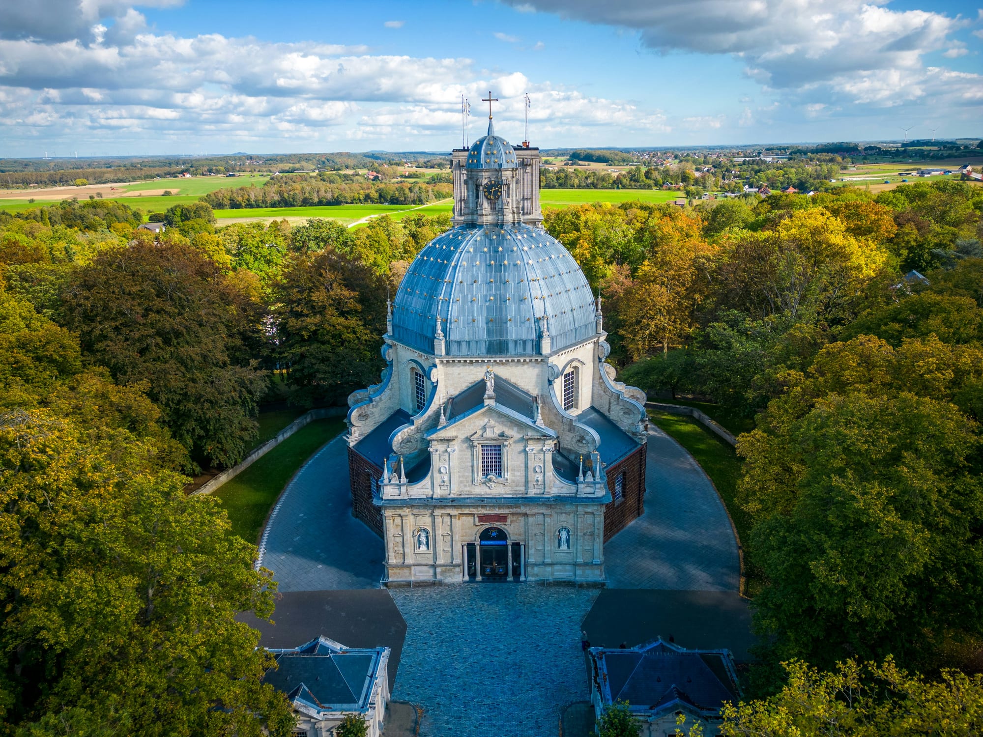 An aerial view of a domed Baroque basilica with a pale grey dome and lantern, set in a ring of autumn trees with farmland stretching to the horizon