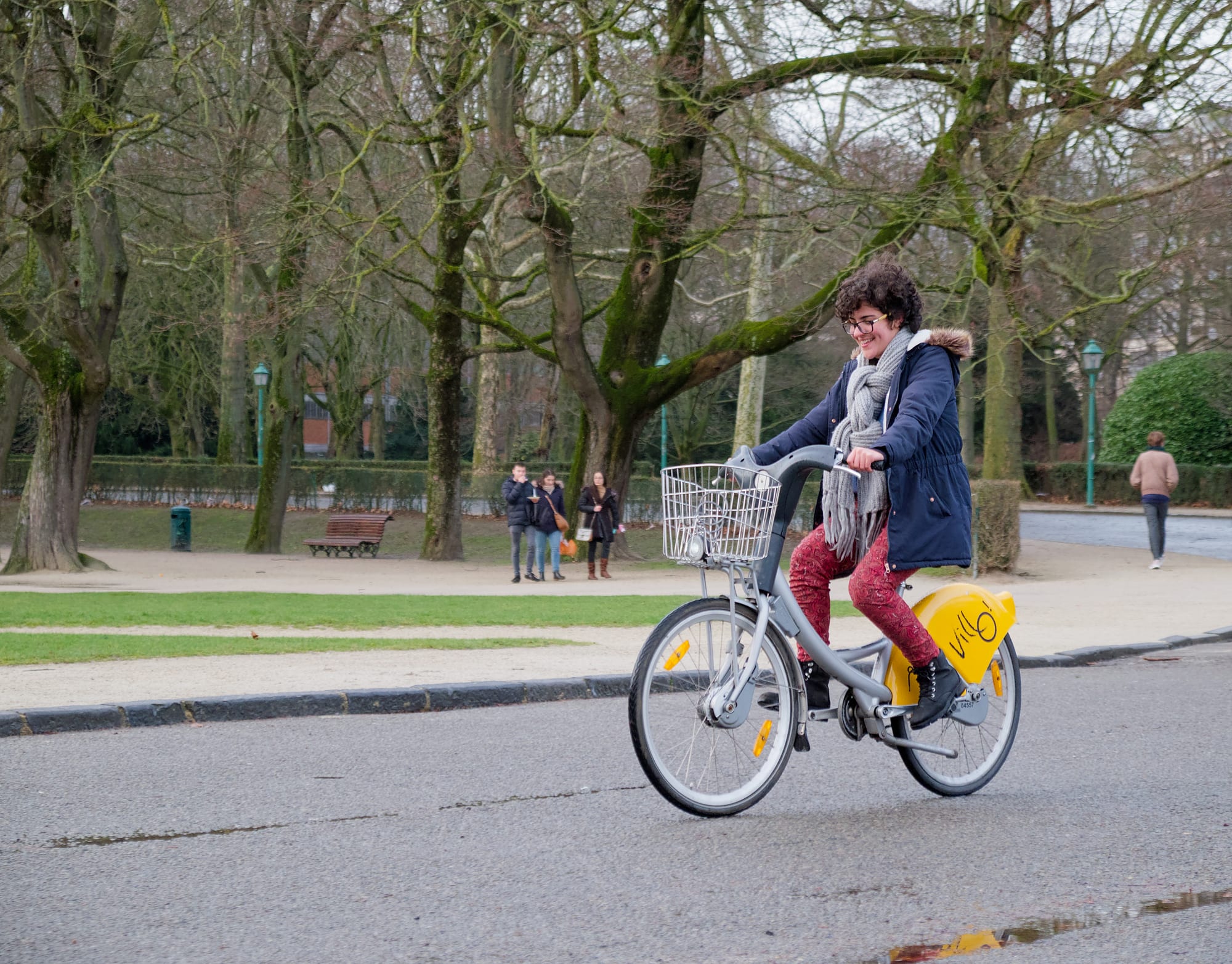 A woman rides a yellow and grey shared bicycle along a paved path in a park, with pedestrians and bare trees behind her