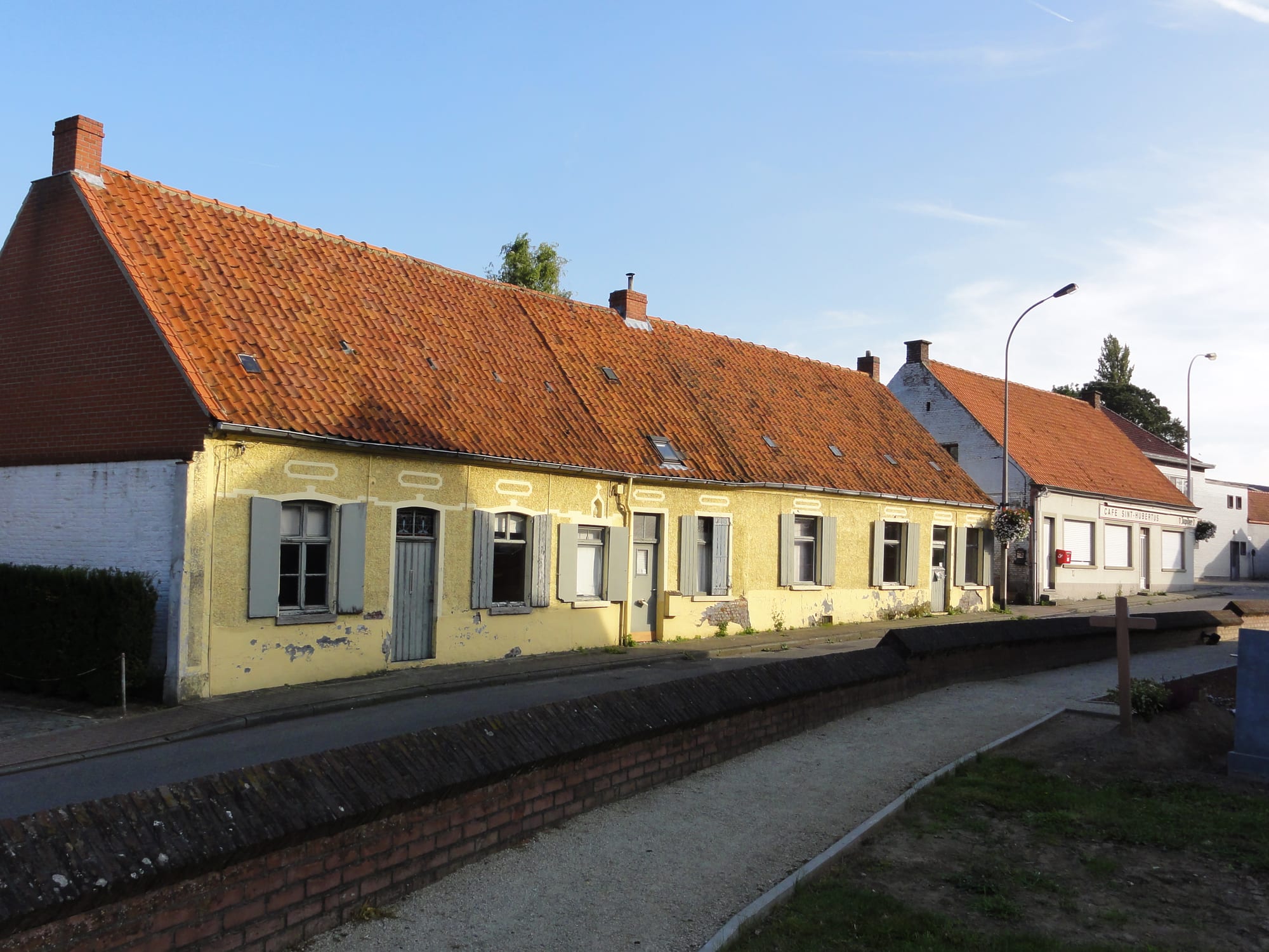 A row of connected single-storey brick houses with tiled roofs and shuttered windows along a village street, a café sign visible further down the road
