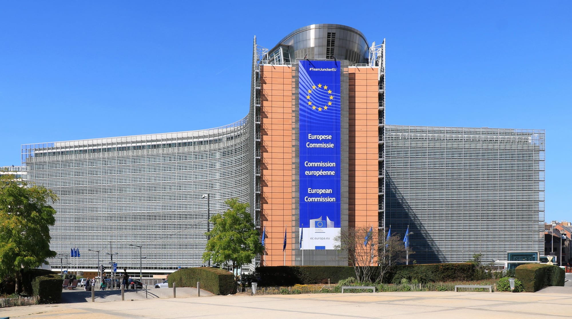 A huge curved office building with a metal-louvred glass facade and a tall blue European Commission banner down its central tower, under a clear sky