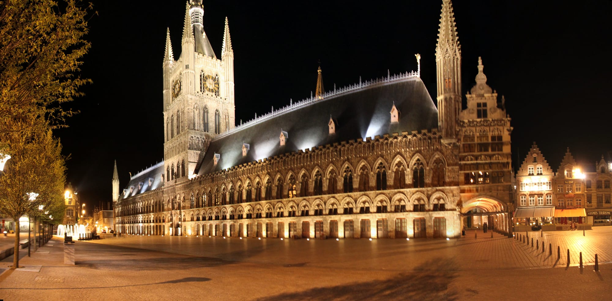 A long floodlit Gothic hall with rows of pointed arches, a steep slate roof and a tall central belfry, on an empty cobbled square at night