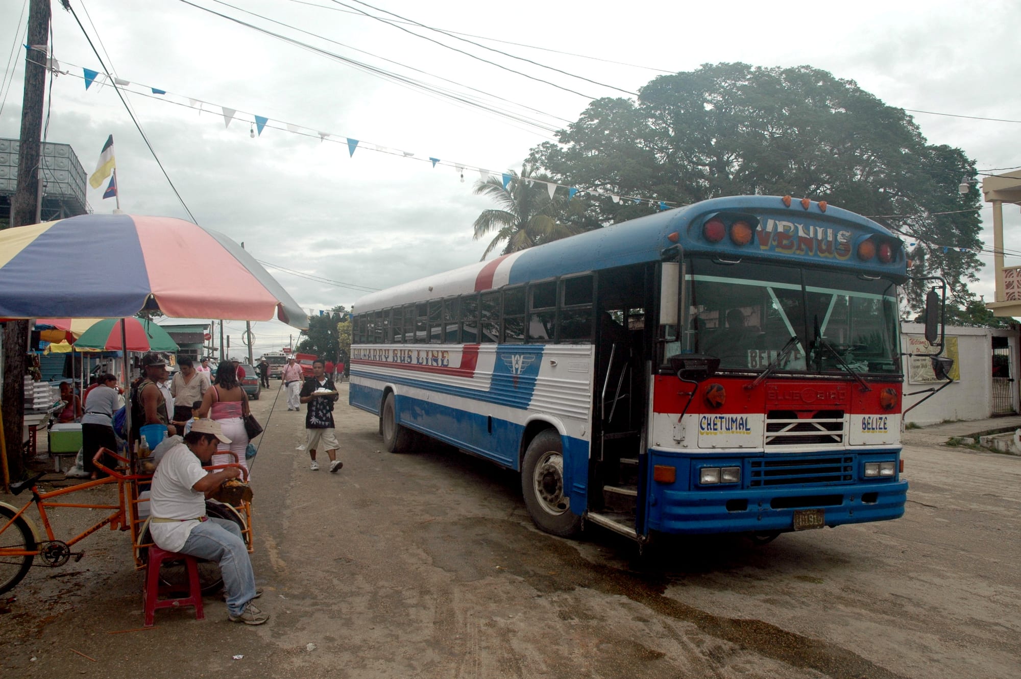 A blue and white bus, formerly an American school bus and repainted for the Gilharry Bus Line, parked on a street in Orange Walk Town beside market umbrellas and pedestrians