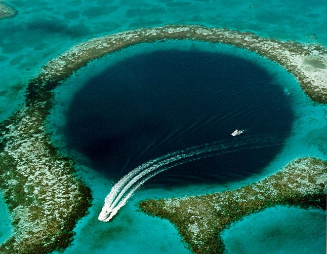Aerial view of a circular dark-blue sinkhole ringed by shallow reef and breaking waves