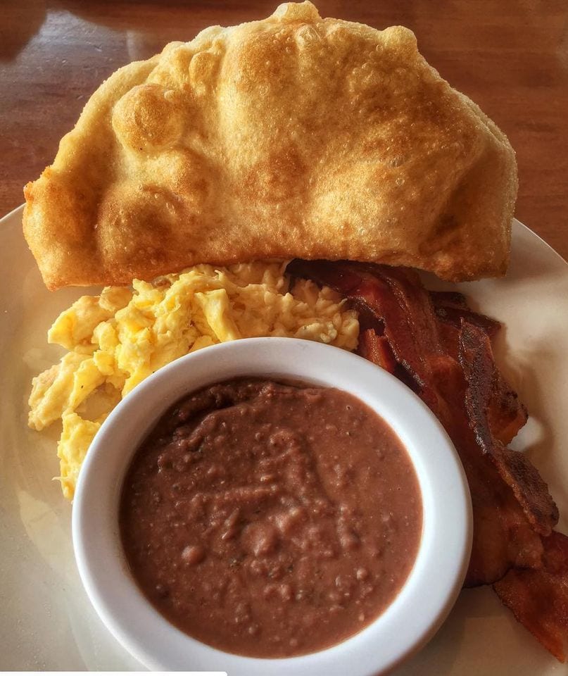 A piece of fried, puffed dough on a plate beside fried eggs, bacon and beans
