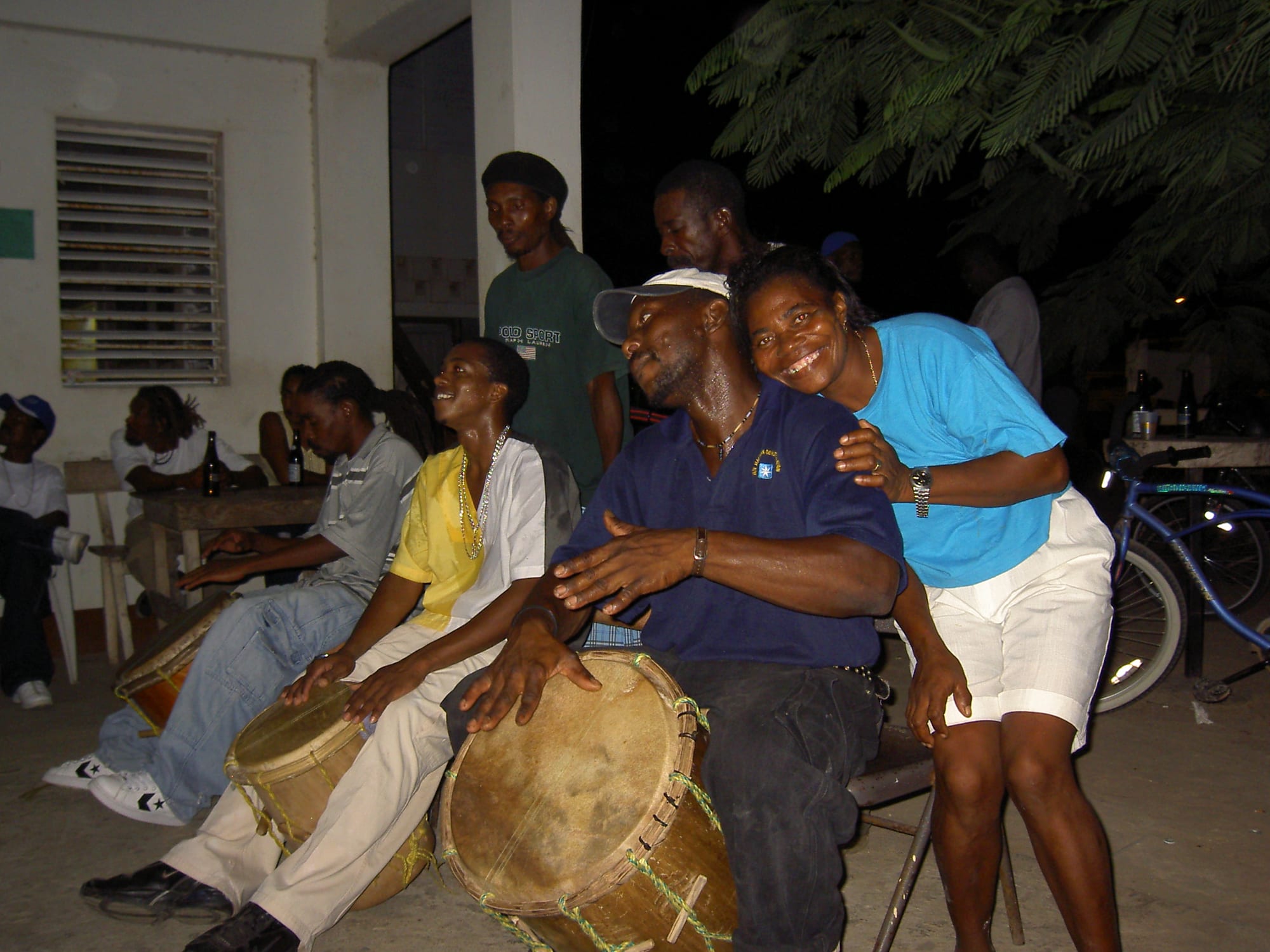 Several men seated in a row playing tall hand drums with laced skins at an evening gathering, while a smiling woman leans on the shoulder of one drummer