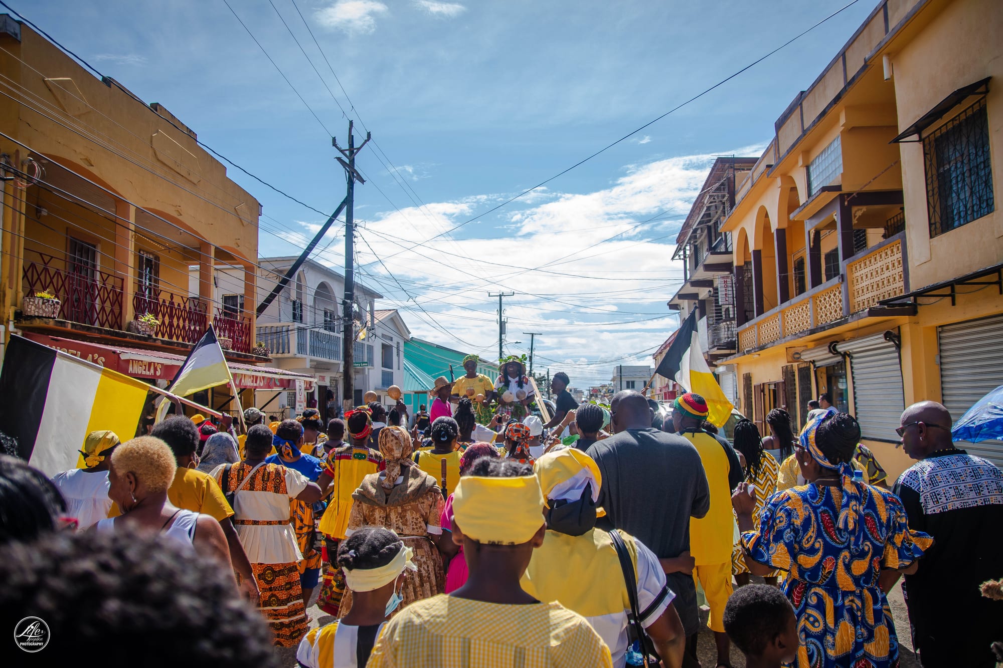 A crowd dressed in yellow, white and patterned African-print clothing walks down a street between colourful two-storey buildings, with black, white and yellow flags held above them