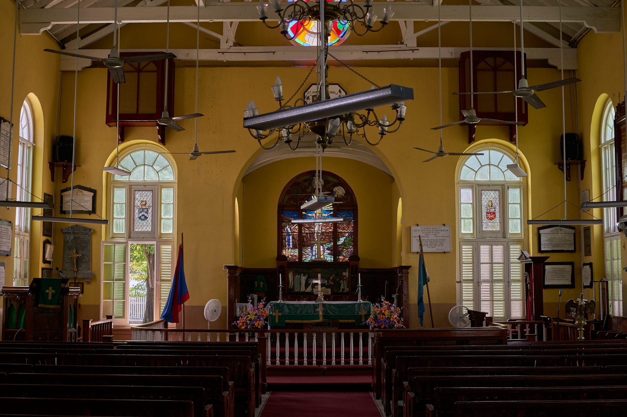 The interior of a church with yellow walls, dark wooden pews, ceiling fans and chandeliers, and a stained-glass window above an altar decorated with flowers