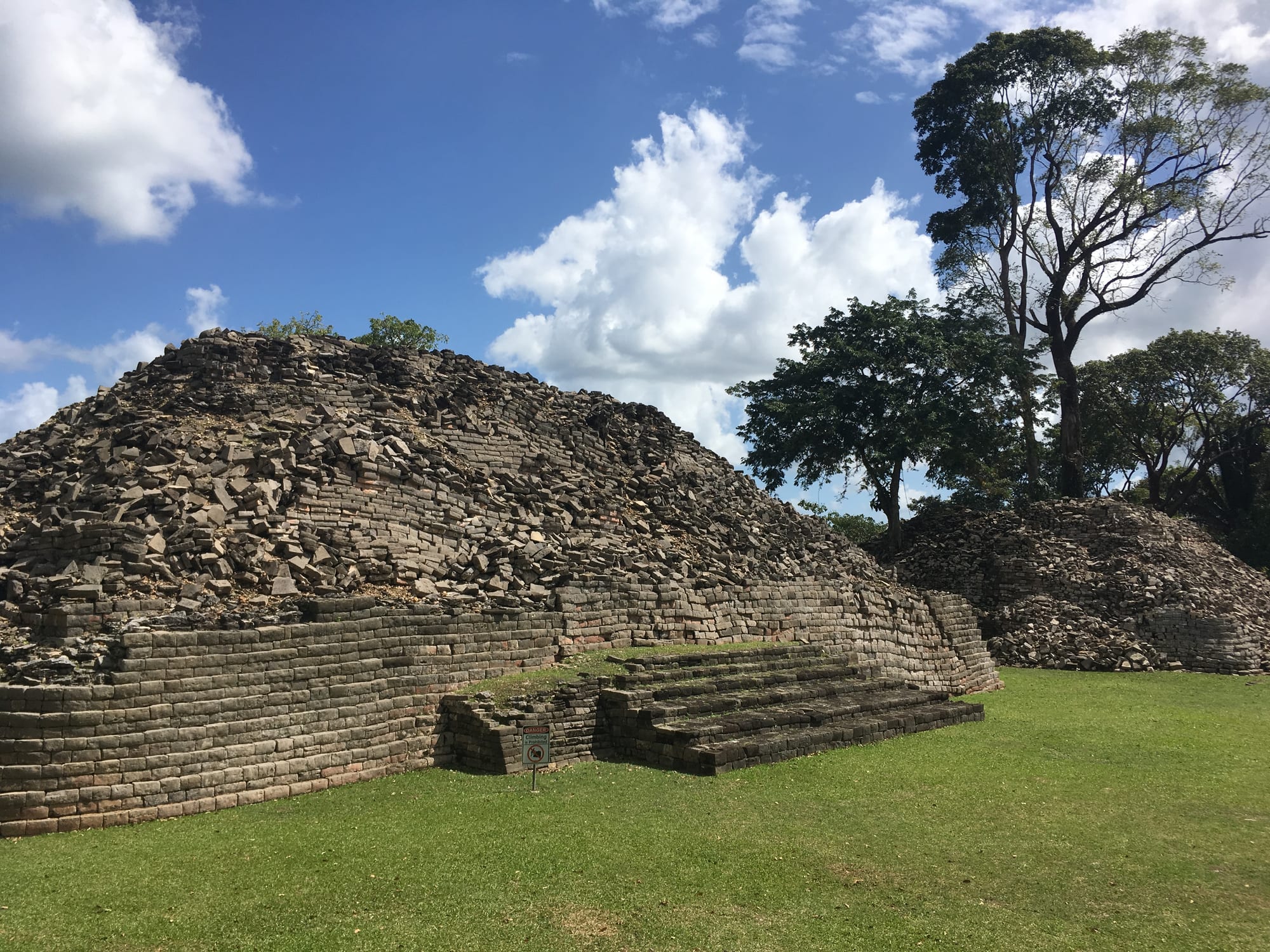 A low Maya platform of dark, finely cut stone blocks with a short stairway, partly collapsed into rubble, on a green lawn with tall trees and white clouds behind