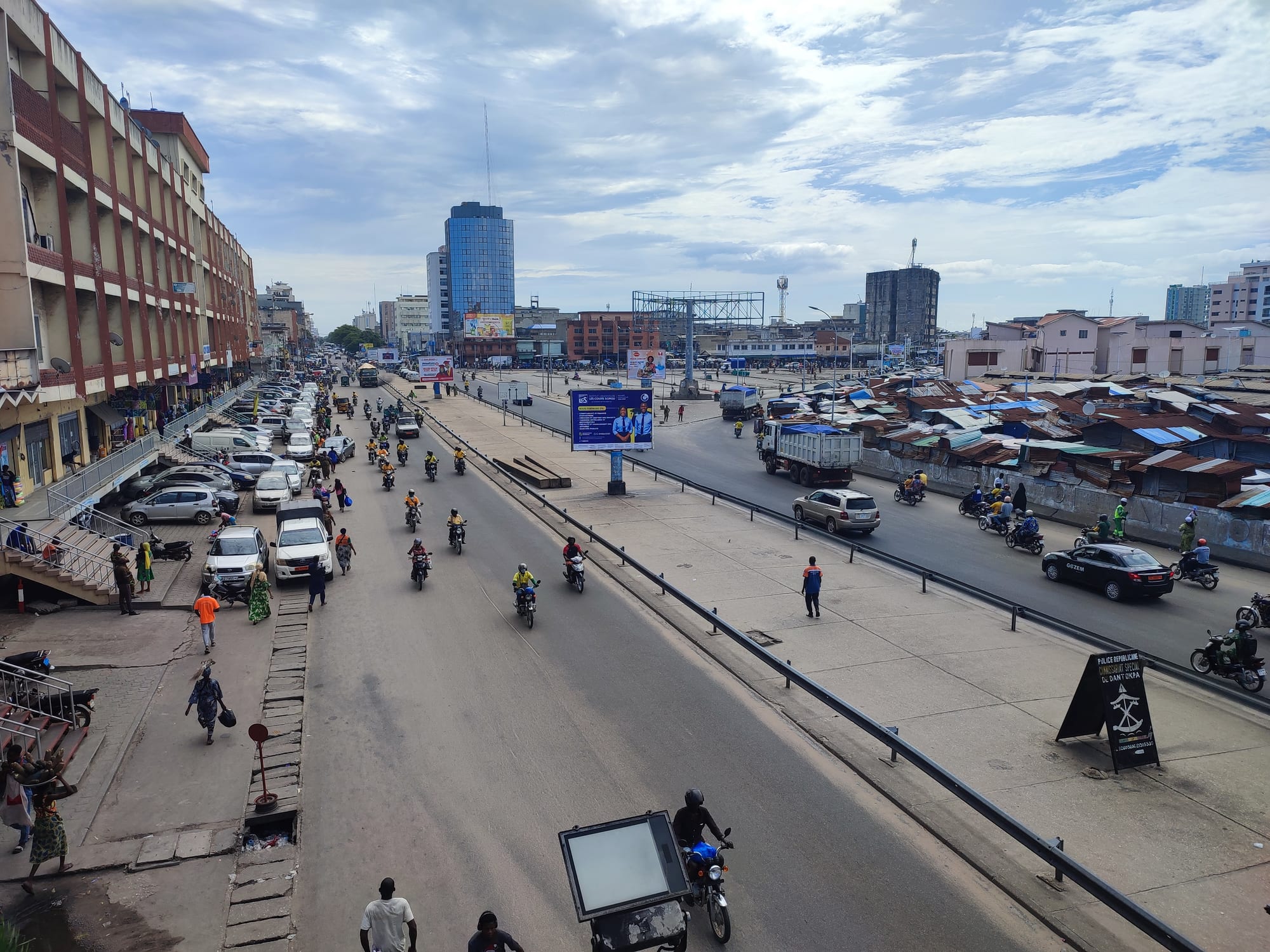 An elevated view of a busy Cotonou street with motorcycle taxis, cars and pedestrians, market rooftops visible in the distance