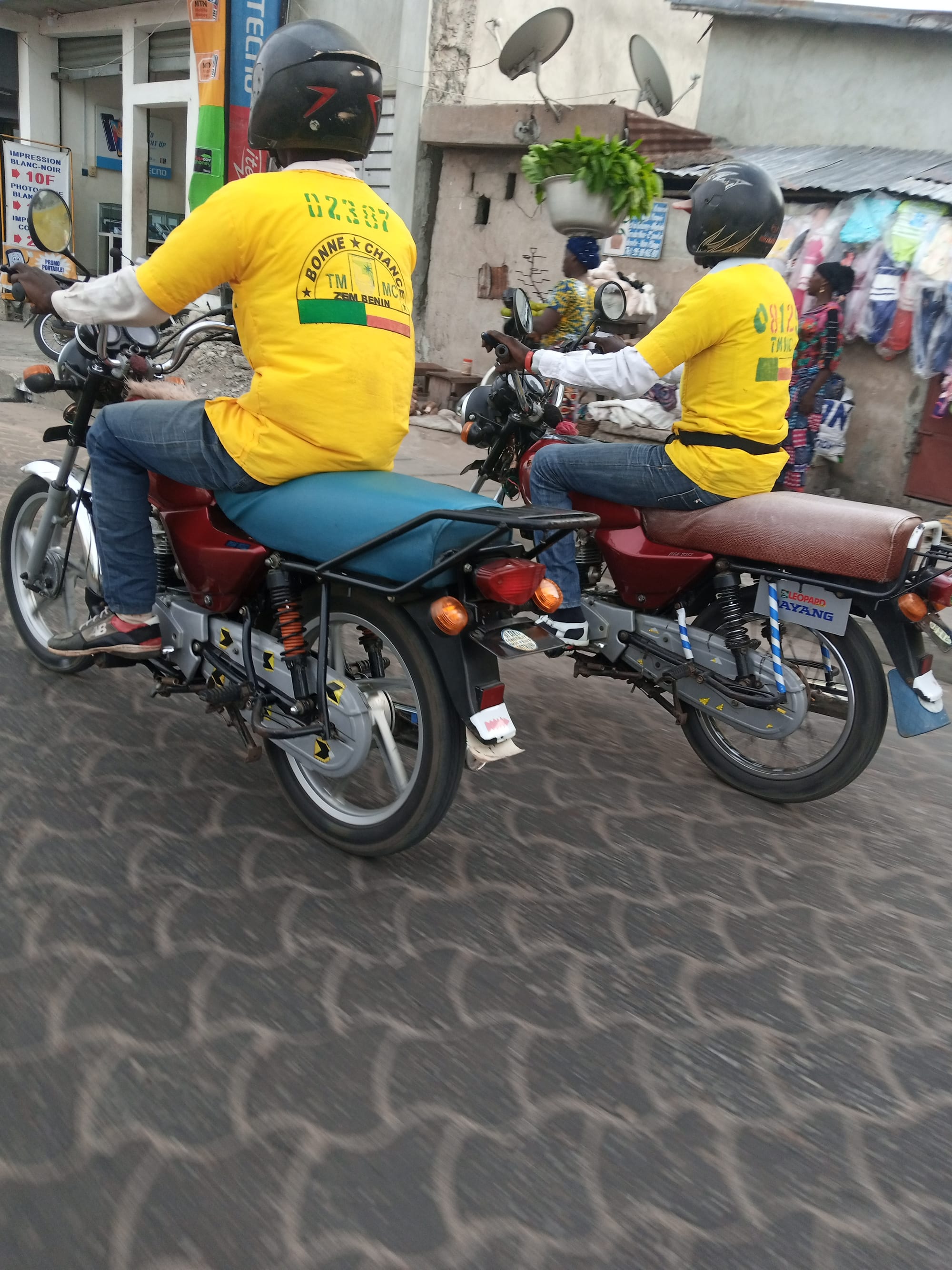 Two motorcycle taxi drivers in matching yellow numbered shirts and helmets riding side by side down a paved street