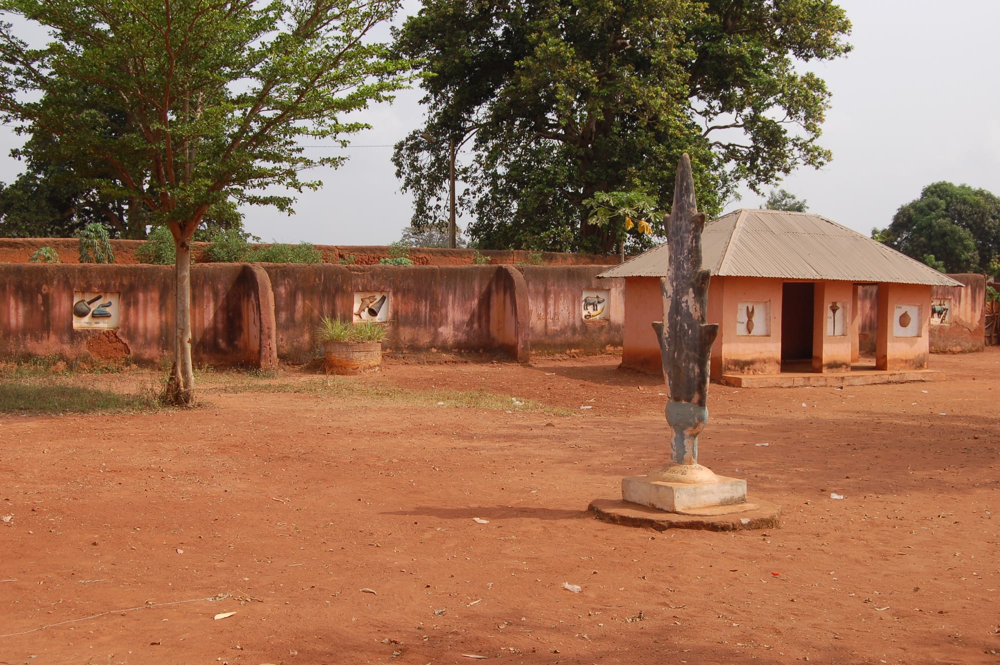 Low reddish mud-walled buildings with thatched and tin roofs behind a bare courtyard