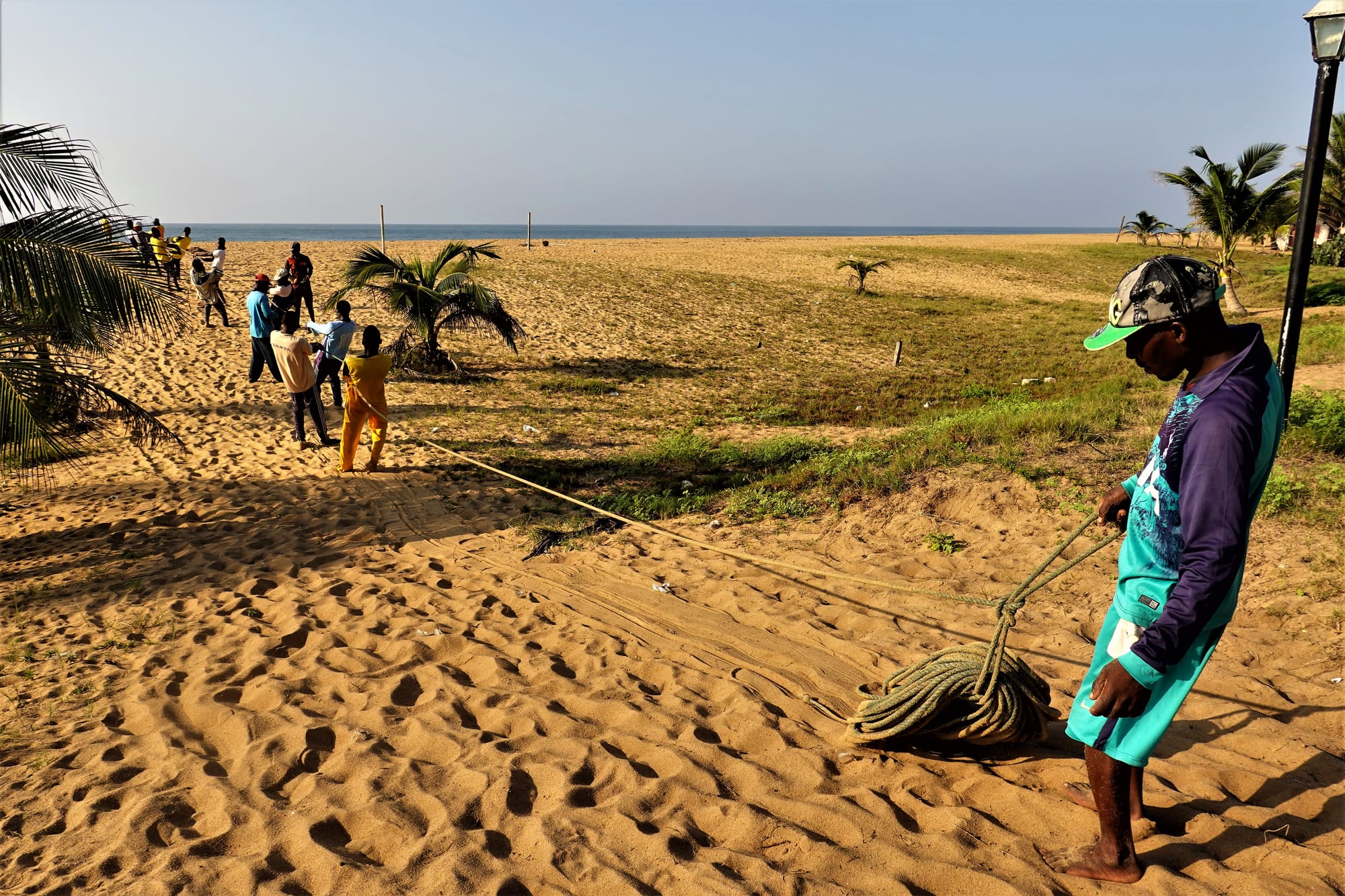 A long line of people hauling a fishing net onto a wide empty beach under a hazy sky