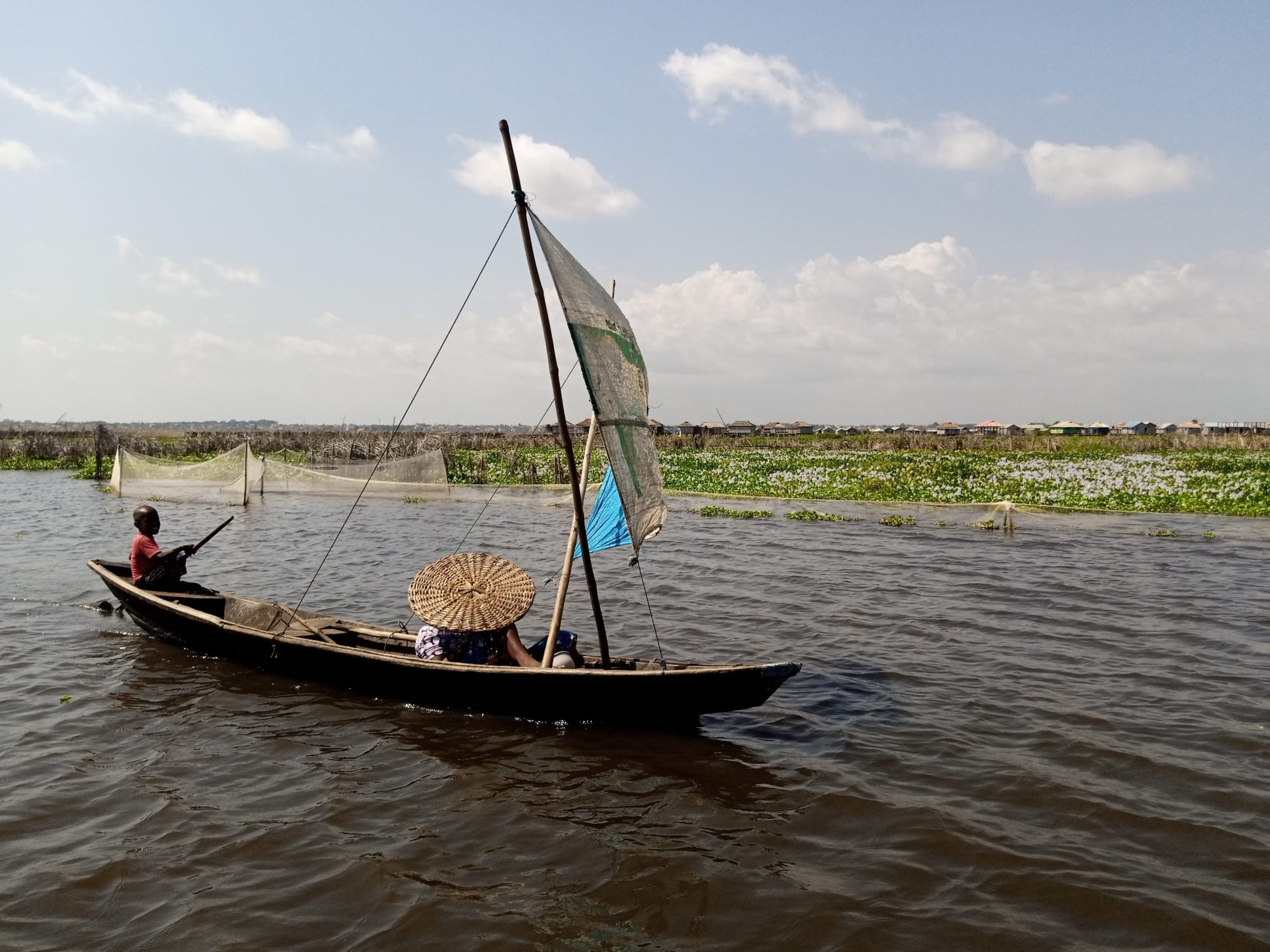 A dugout canoe with a patchwork sail on calm lake water near stilt houses