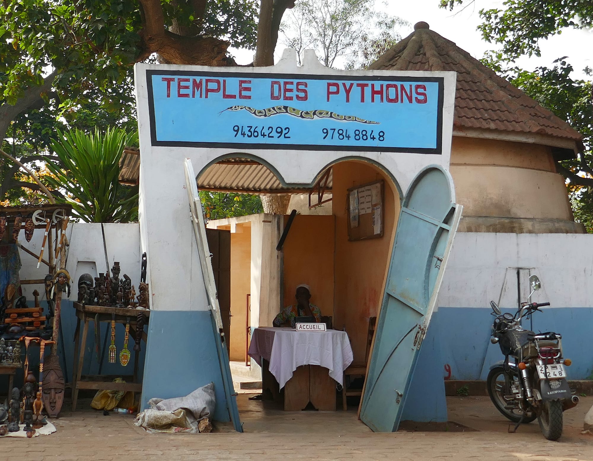 A low pink shrine building with a python relief over the door, in a dusty compound