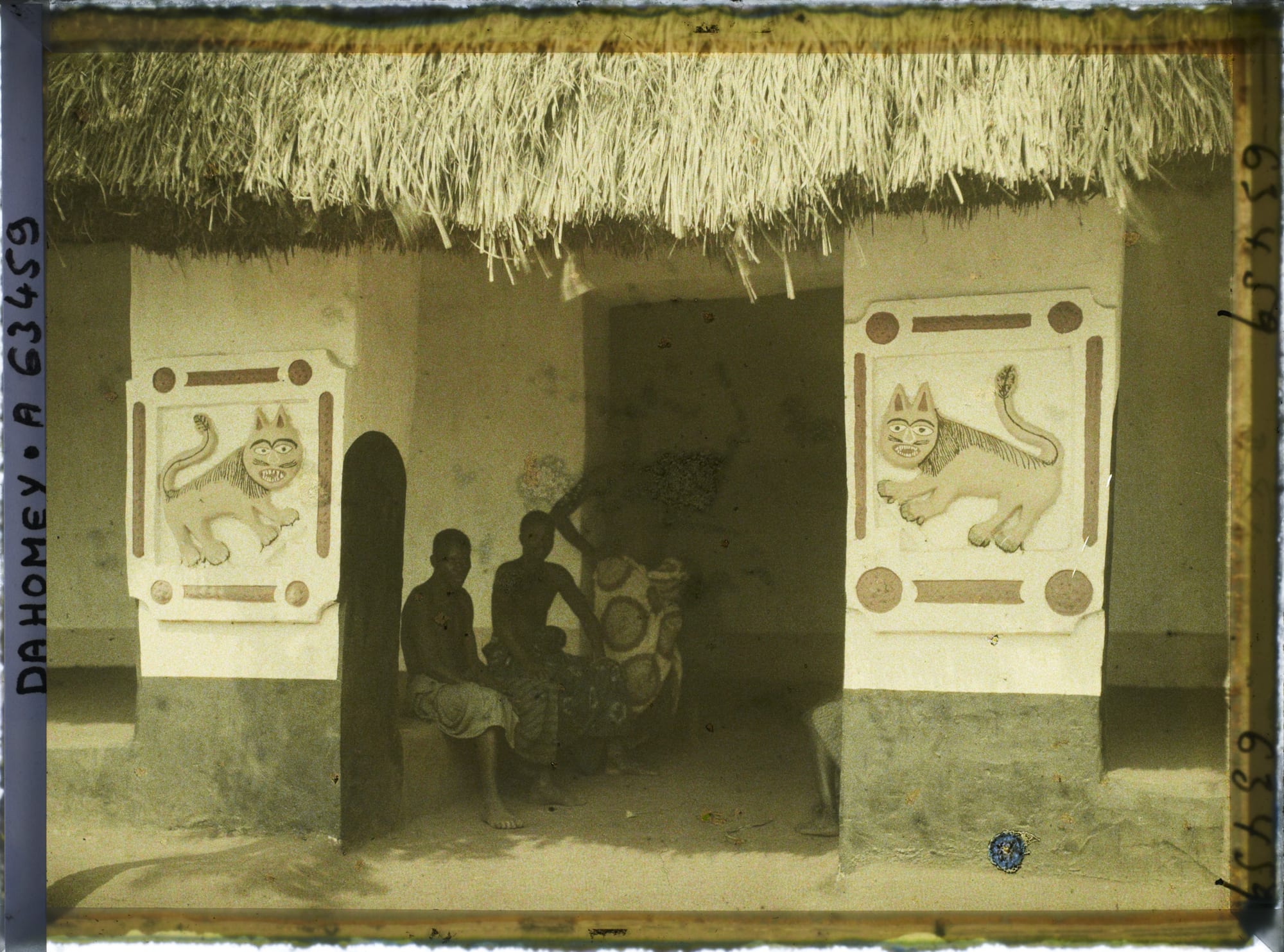 An early photograph of a low mud-walled palace gateway with a thatched roof and figures standing before it