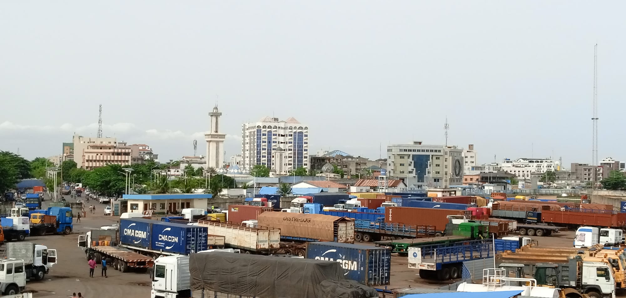 A dense low-rise African city seen from above, with rusted roofs, a mosque, and a lagoon in the distance