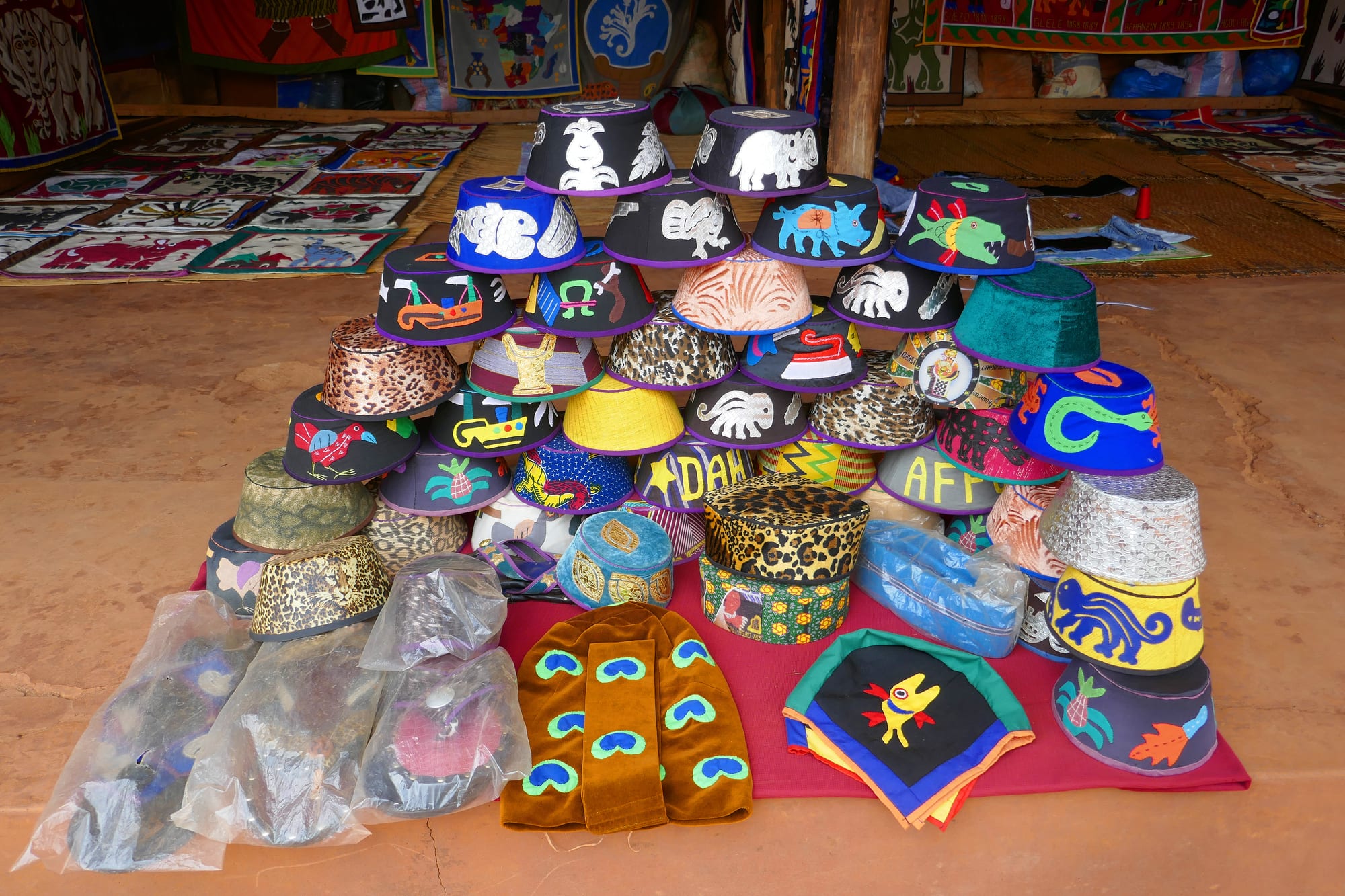 A pyramid of brightly coloured fabric caps decorated with appliqué animals, fish, birds and lettering, set on a red cloth with appliqué wall hangings displayed behind