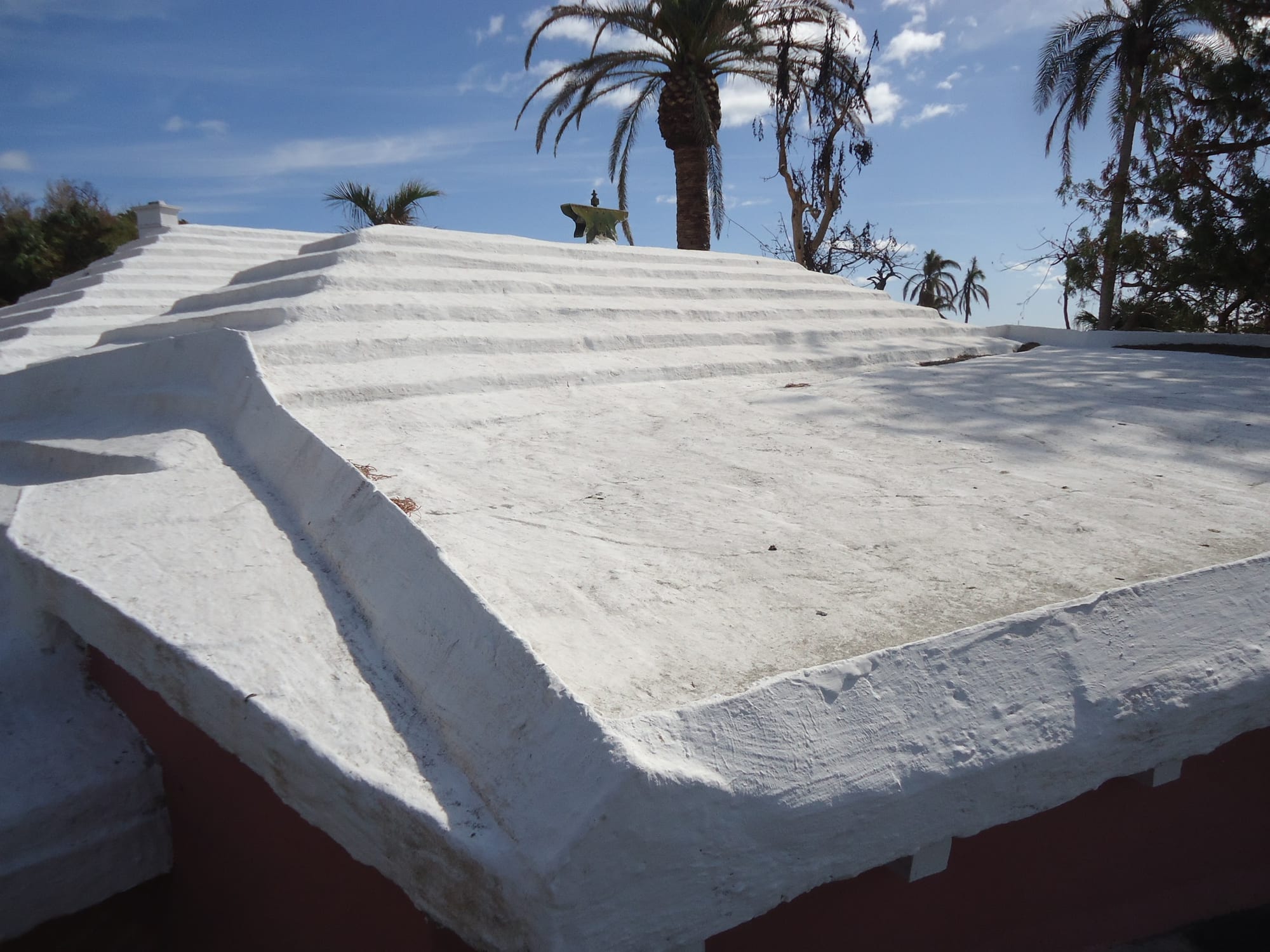 A close view of a white-painted, stepped limestone roof under a blue sky with palm trees behind