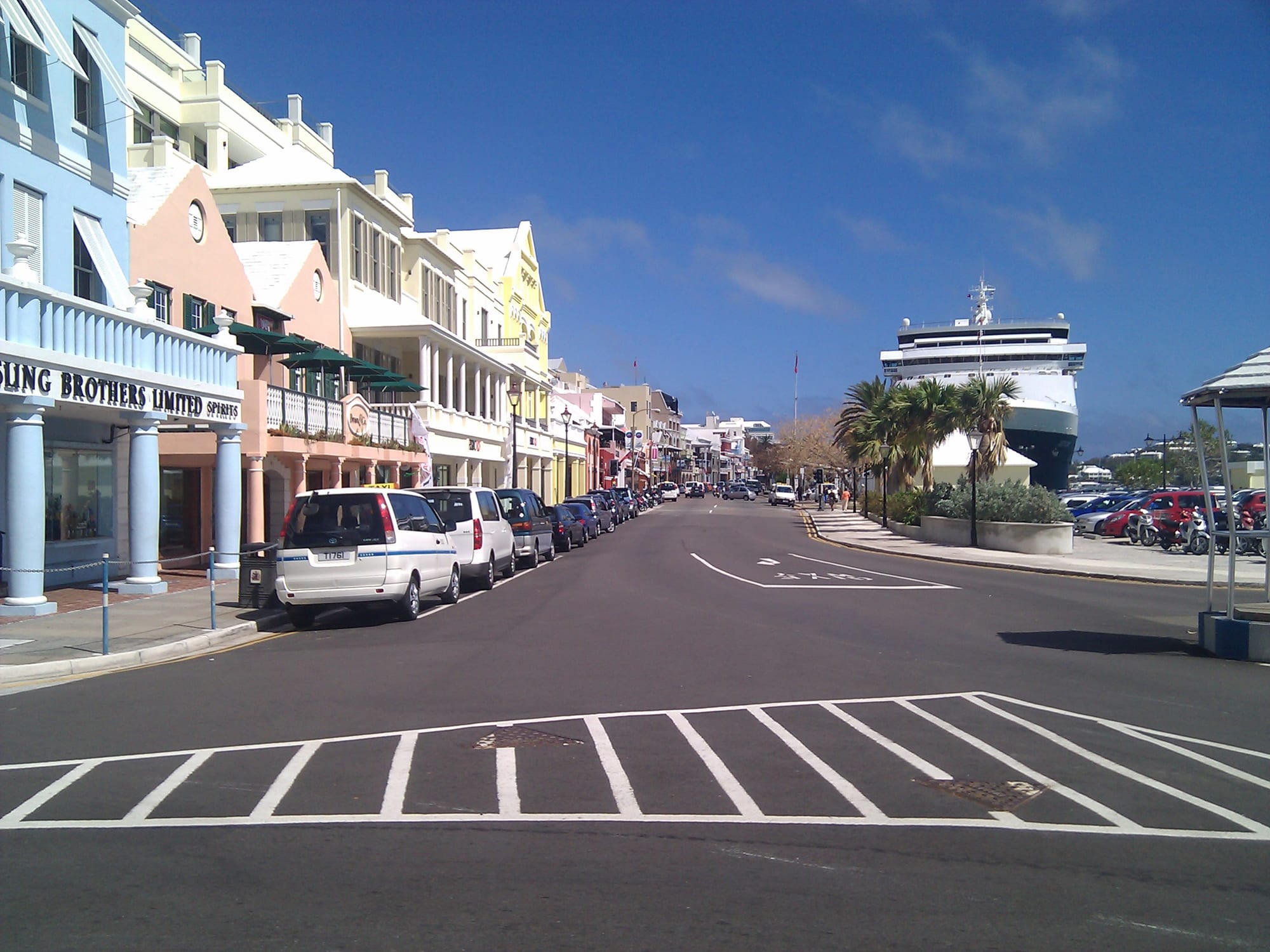 A sunlit view down Front Street in Hamilton, Bermuda, lined with pastel-painted colonial buildings, parked cars and palm trees, with a cruise ship docked at the far end
