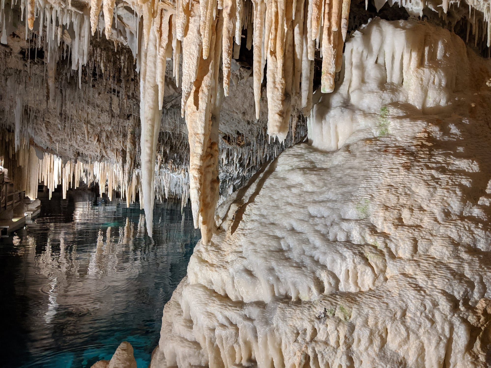 A cavern of pale stalactites reflected in a still, glass-clear underground pool crossed by a floating walkway