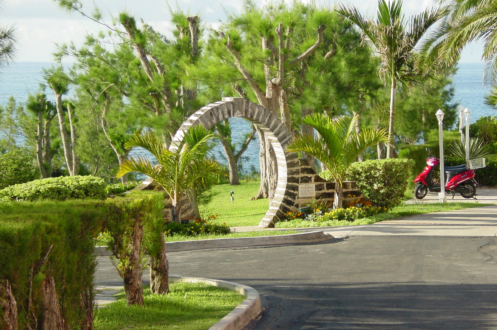 A circular arch of pale stone standing on a lawn among palms and casuarina trees, with the blue sea visible beyond
