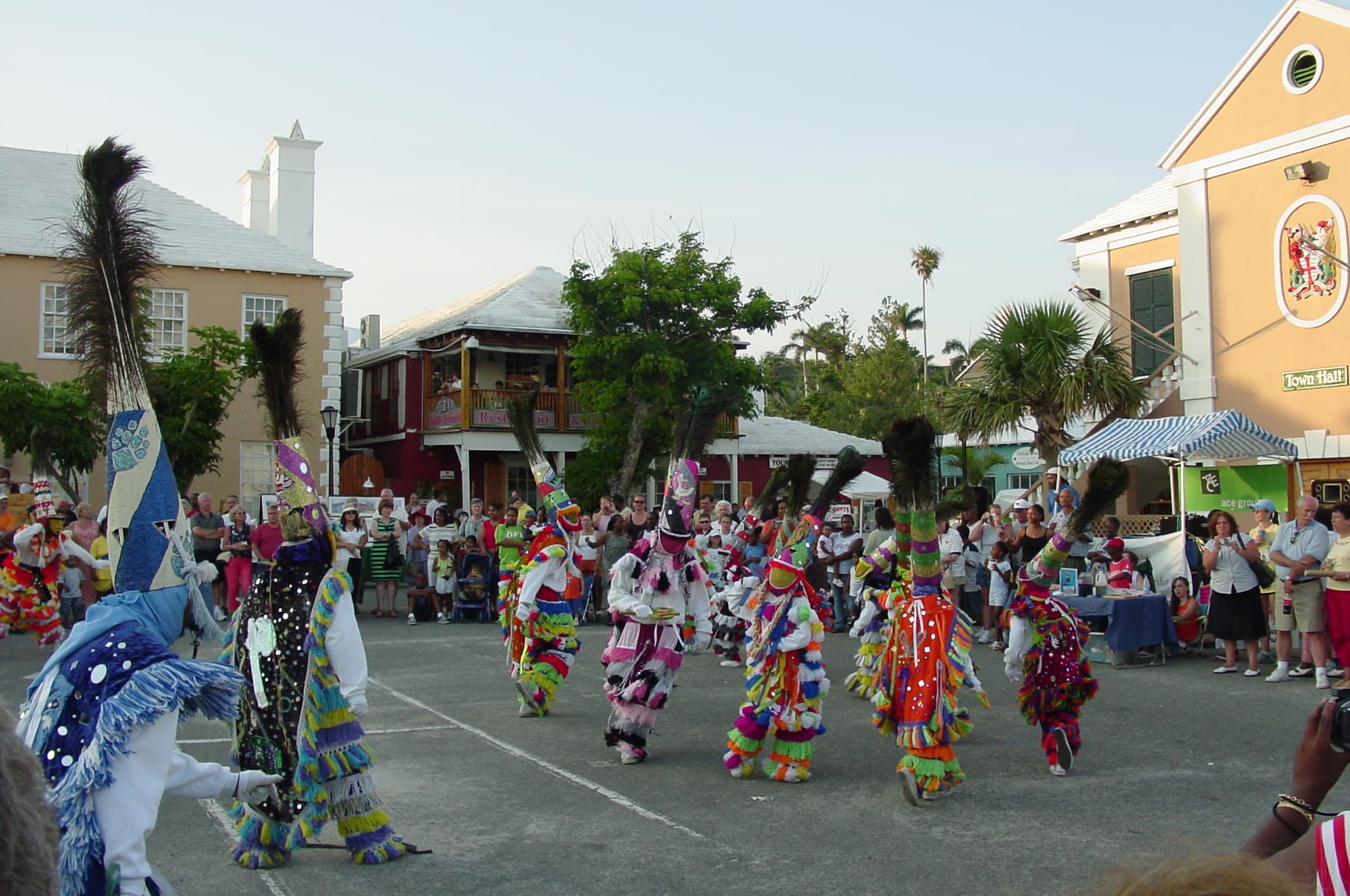 A troupe of Gombey dancers in masks, brightly fringed costumes and tall headdresses topped with peacock feathers dance across a paved square, watched by a crowd in front of pastel-coloured buildings with white roofs and palm trees