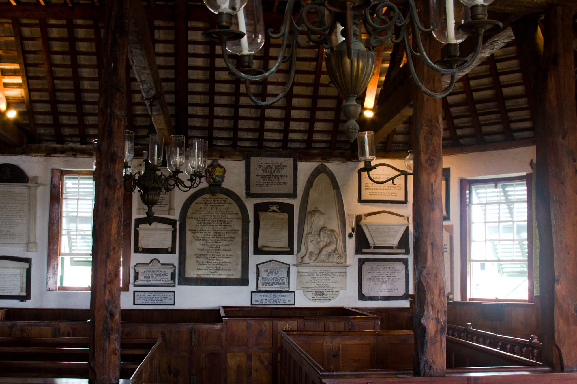 The interior of an old church with whitewashed walls covered in carved and painted memorial tablets, dark wooden box pews, exposed timber roof beams and hanging brass candle chandeliers