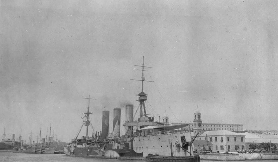 An early 20th-century battleship moored at a stone naval dockyard with cranes and workshops