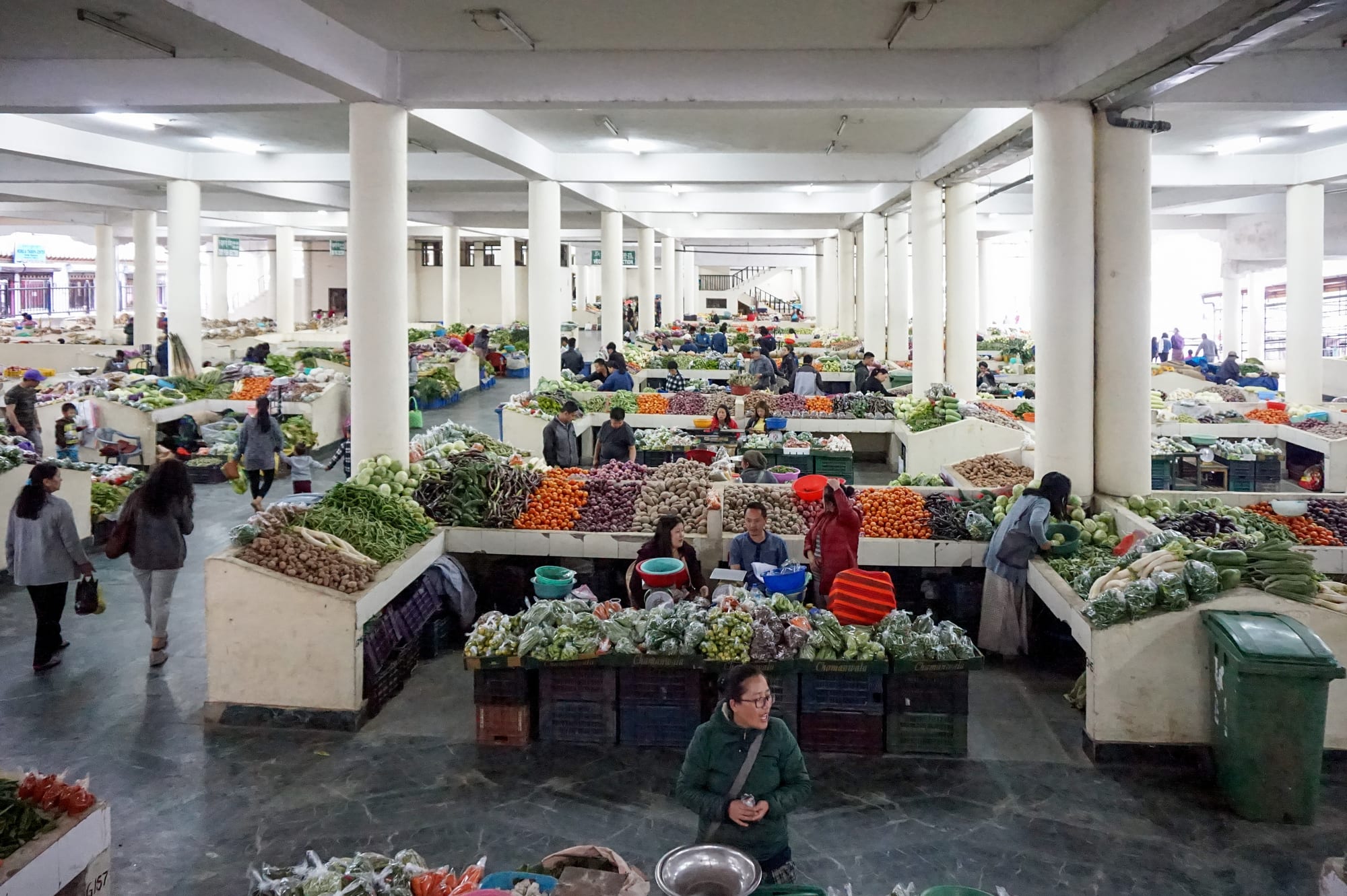 Rows of vegetable stalls piled with produce inside a large covered market hall, with shoppers browsing between the stands