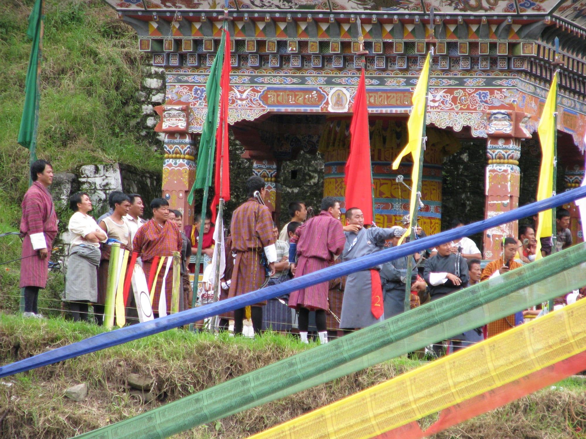 A group of men in traditional striped robes gathered near a decorated wooden pavilion with colourful flags, at an archery event