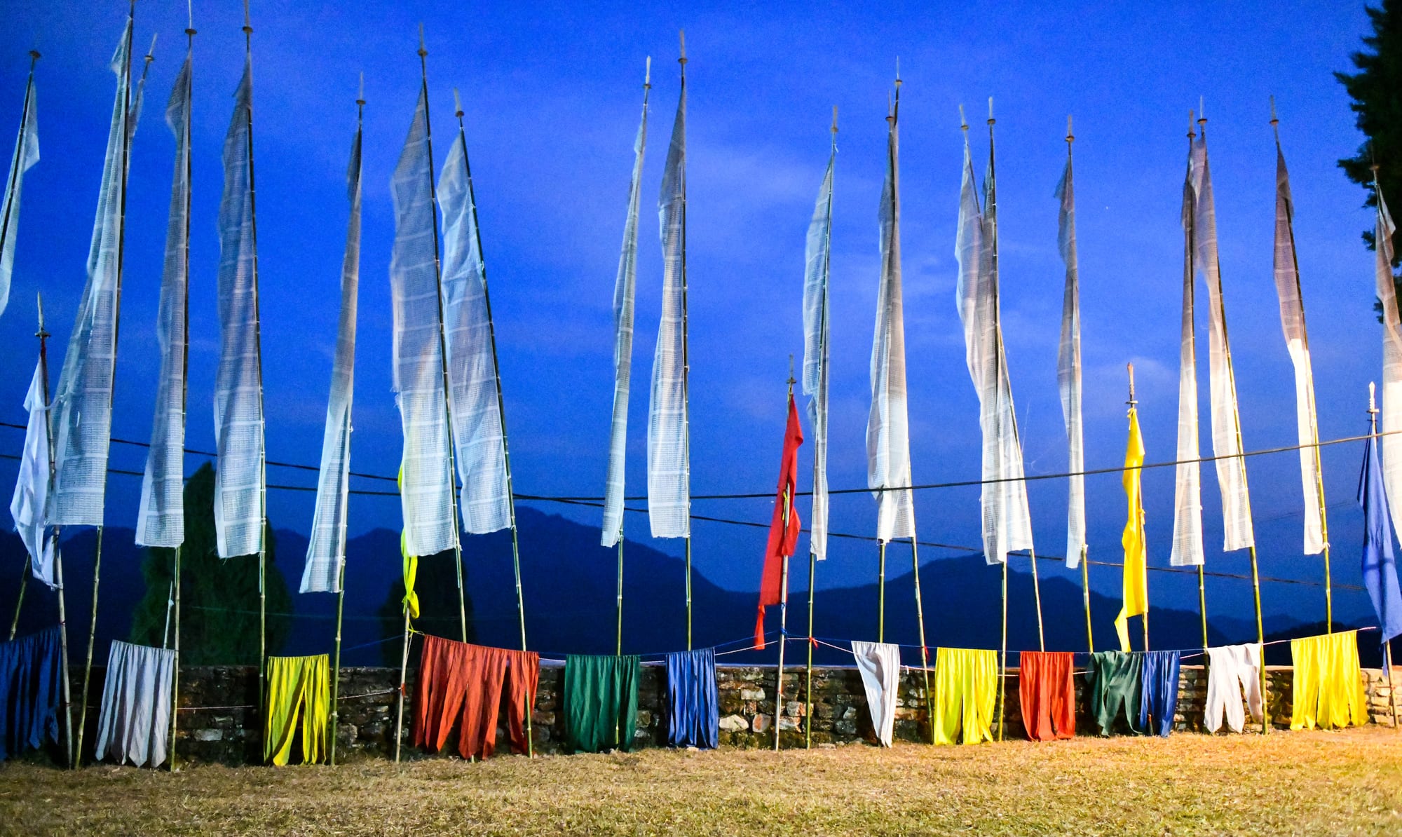 A row of tall white prayer flags on long bamboo poles stands against a deep blue evening sky, with smaller red, yellow, green and blue cloths hung along a low stone wall beneath them and mountains in the distance