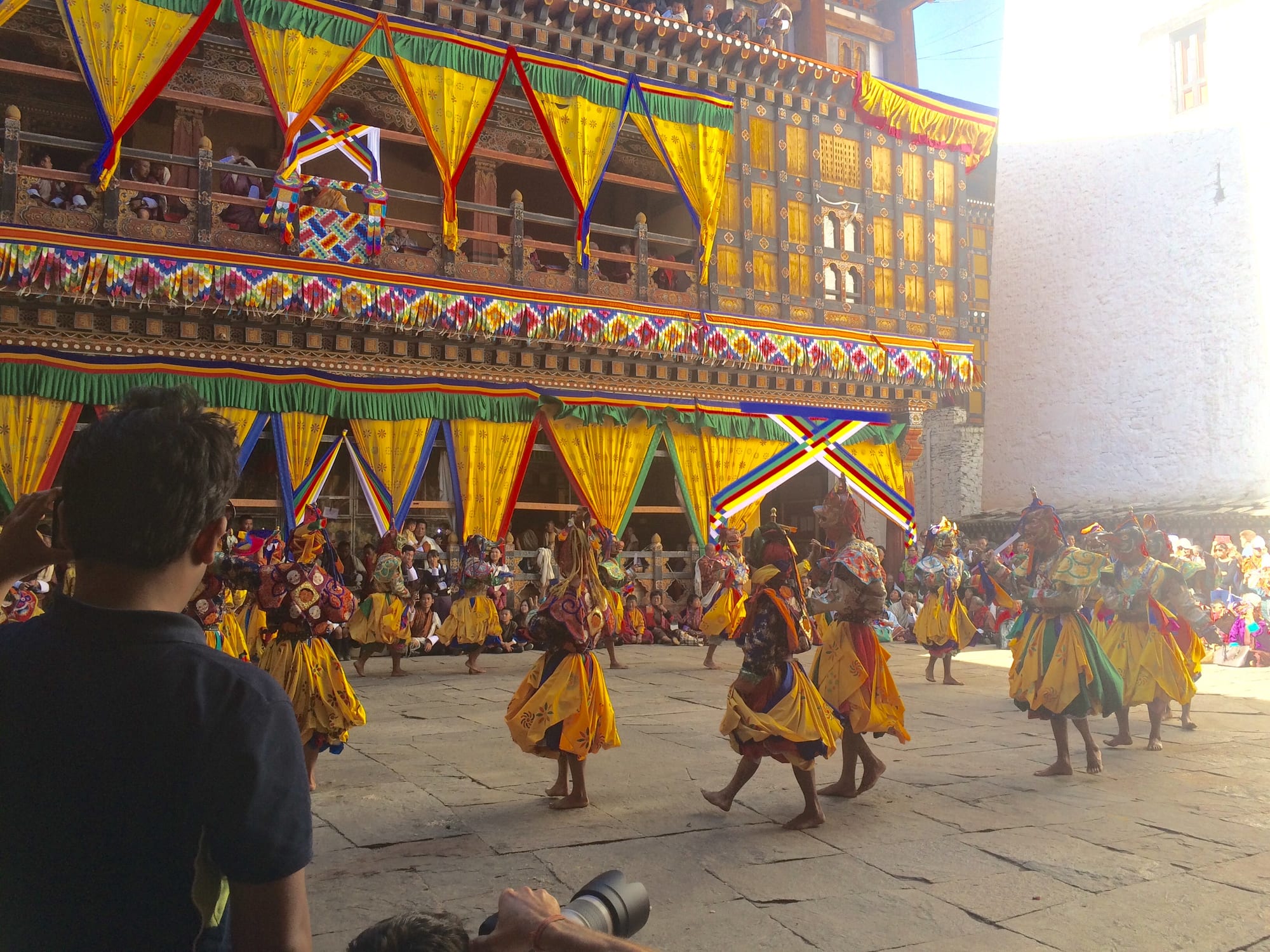 Masked dancers in flowing yellow and multicoloured costumes whirl across a stone courtyard in front of a monastery building hung with bright yellow, red and blue drapes, watched by crowds seated around the edge and on the balconies