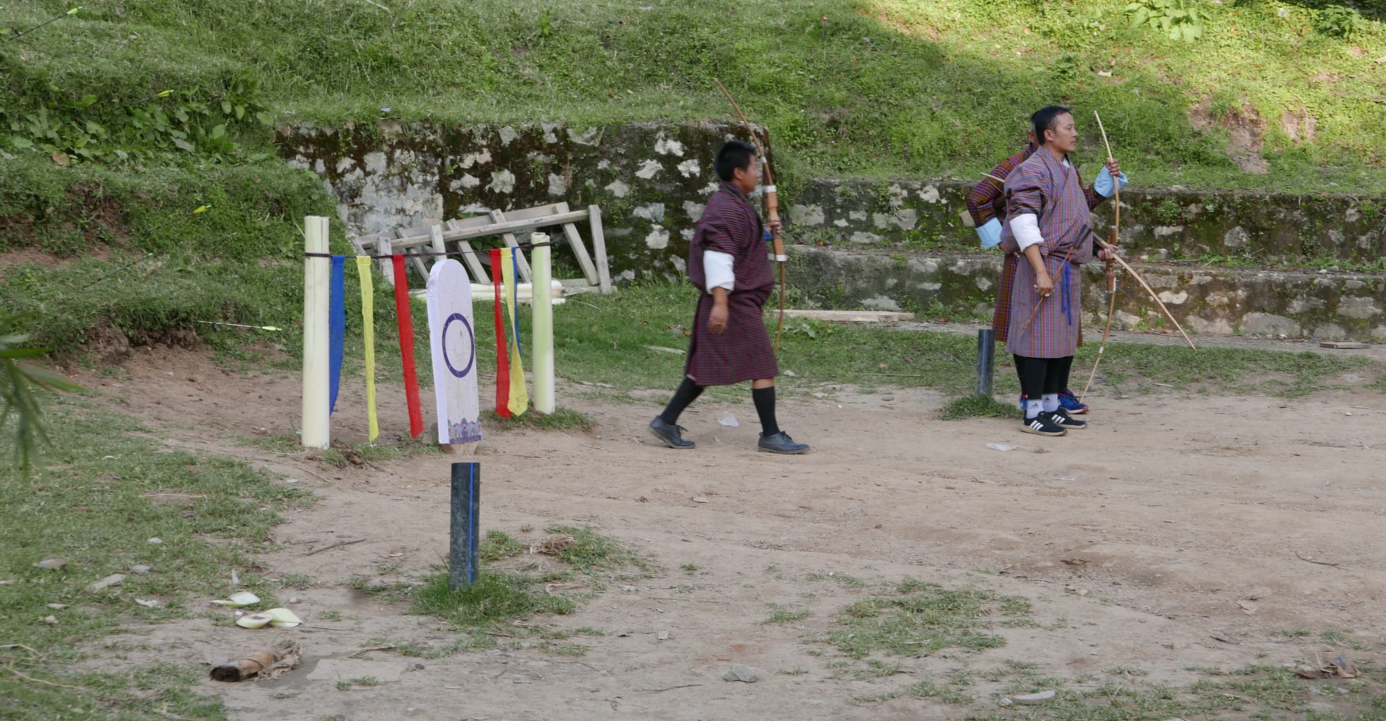 Three archers in checked gho robes and knee socks on a dusty archery range, one holding a traditional bamboo bow and two holding compound bows, beside a small painted target and coloured flags on poles