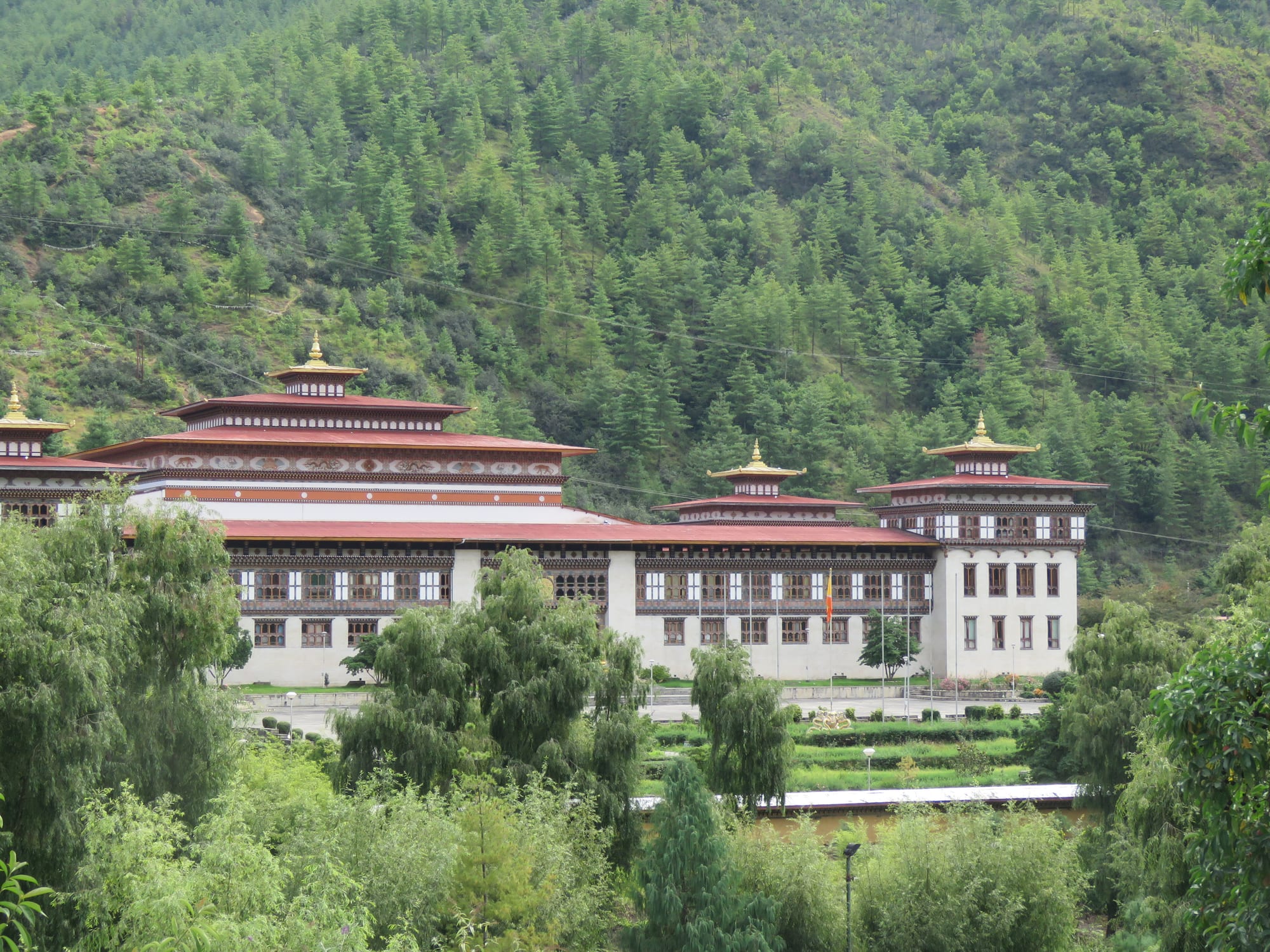A long white building with rows of timber windows and tiered red roofs topped by golden finials, set among willows below a pine-covered hillside
