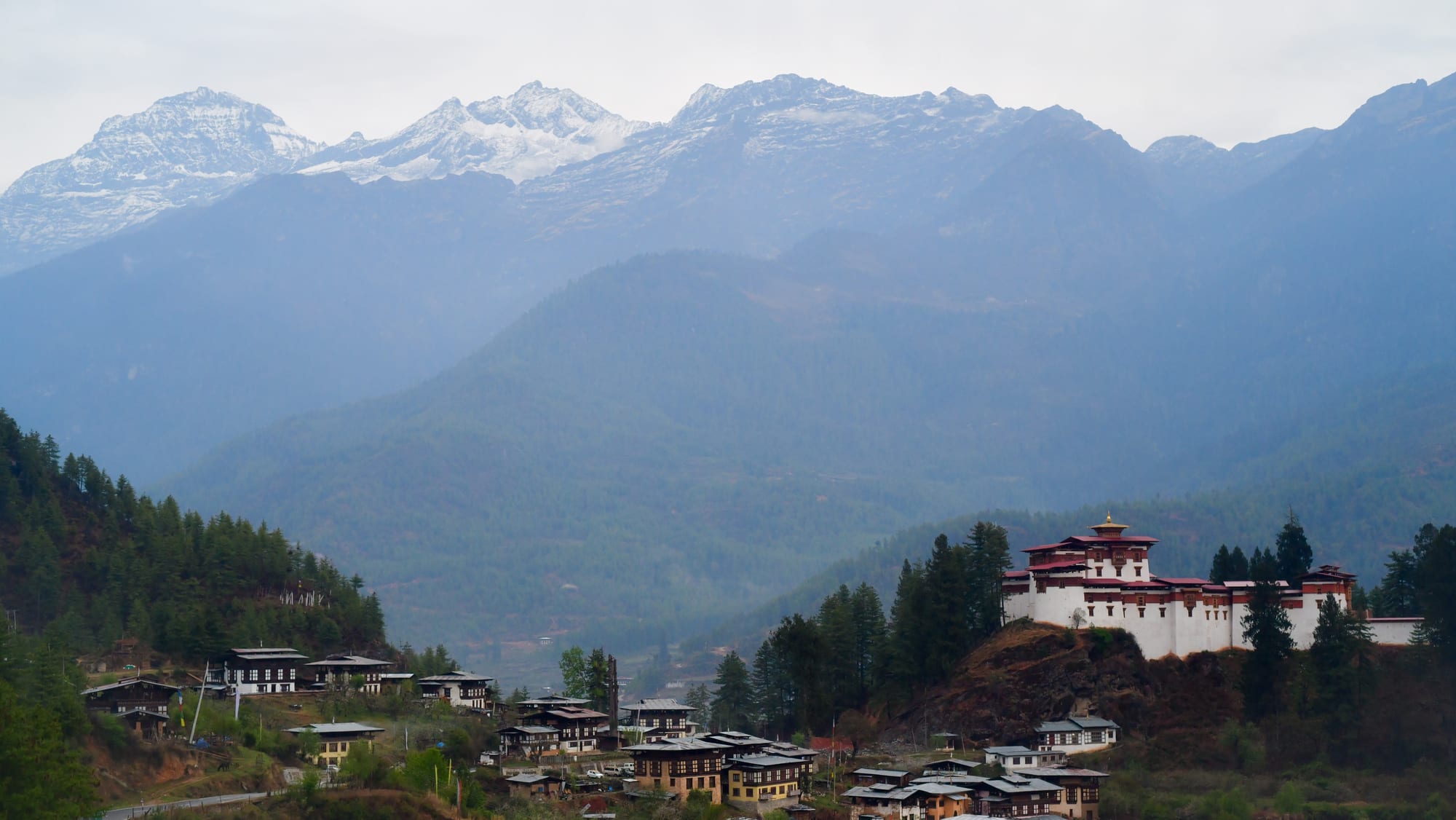 A white fortress with red-banded walls on a rocky knoll above a village, with forested ridges and snow-capped Himalayan peaks behind