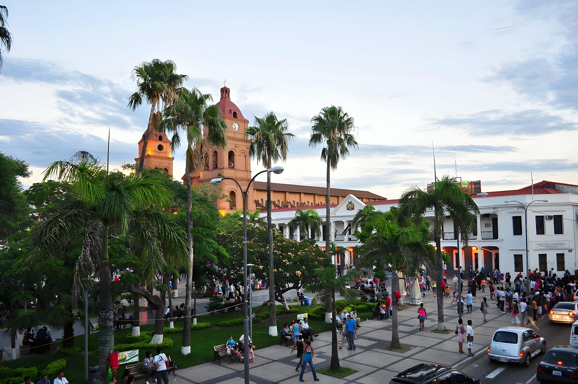 Families and couples walking and sitting on benches in a palm-lined plaza in Santa Cruz de la Sierra at dusk, a colonial-era cathedral behind
