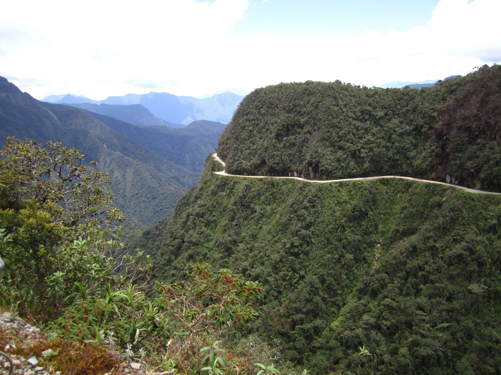 A narrow gravel road with a sheer unguarded drop on one side, cut into a green cliff wrapped in cloud