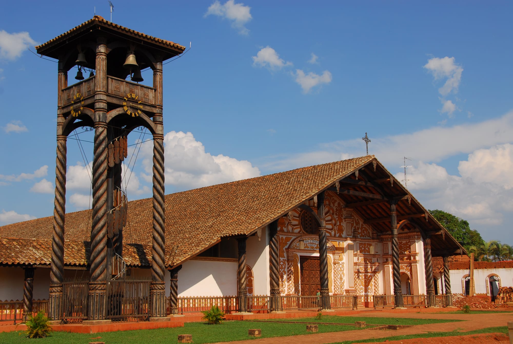 A grand Baroque mission church with a wooden bell tower and carved facade, jungle beyond