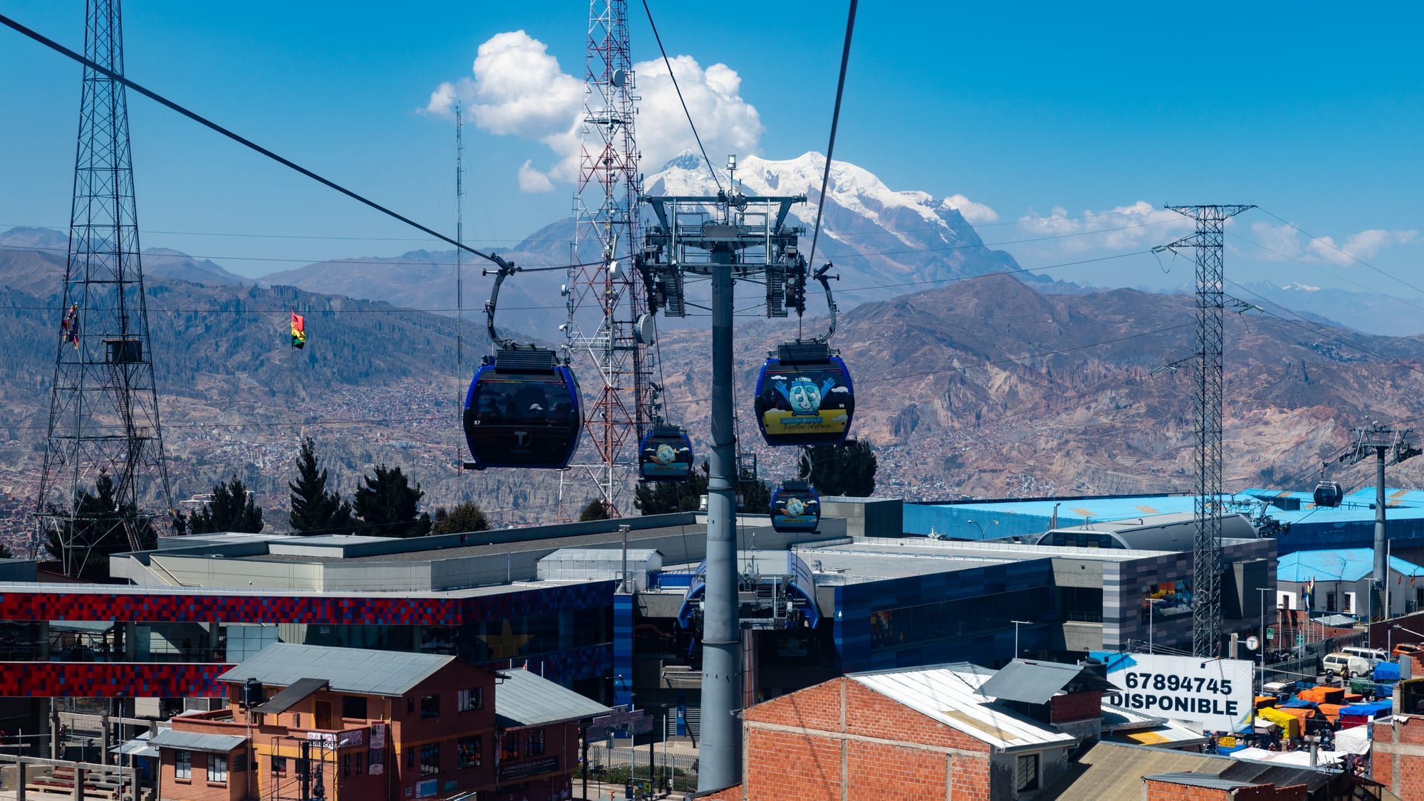 Gondola cable cars crossing above a dense city of brick buildings, a huge snow peak behind
