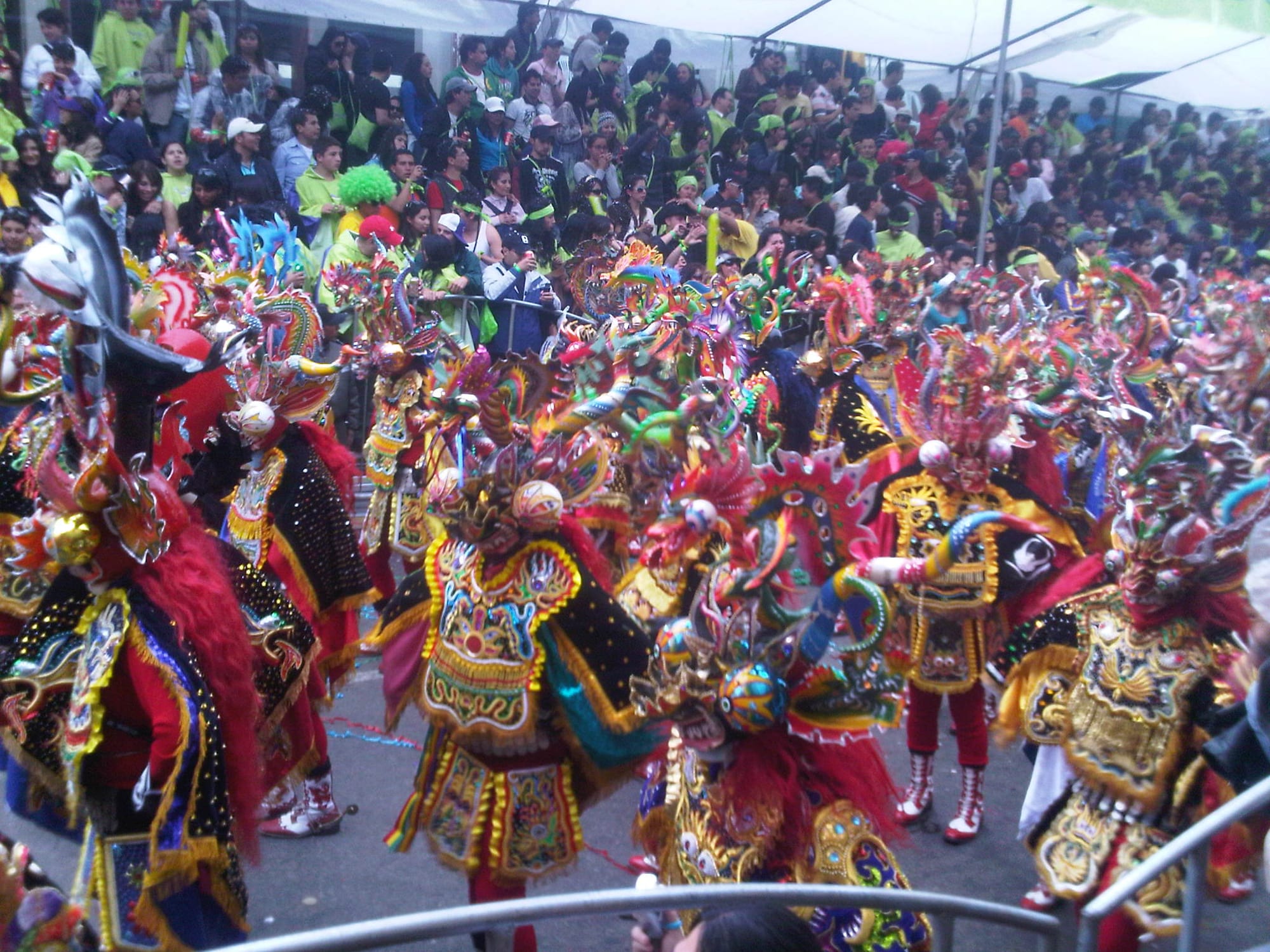 A mass of dancers in ornate devil costumes with horned, dragon-like masks, embroidered capes in gold, red and black and tall boots moves along a street past packed spectator stands