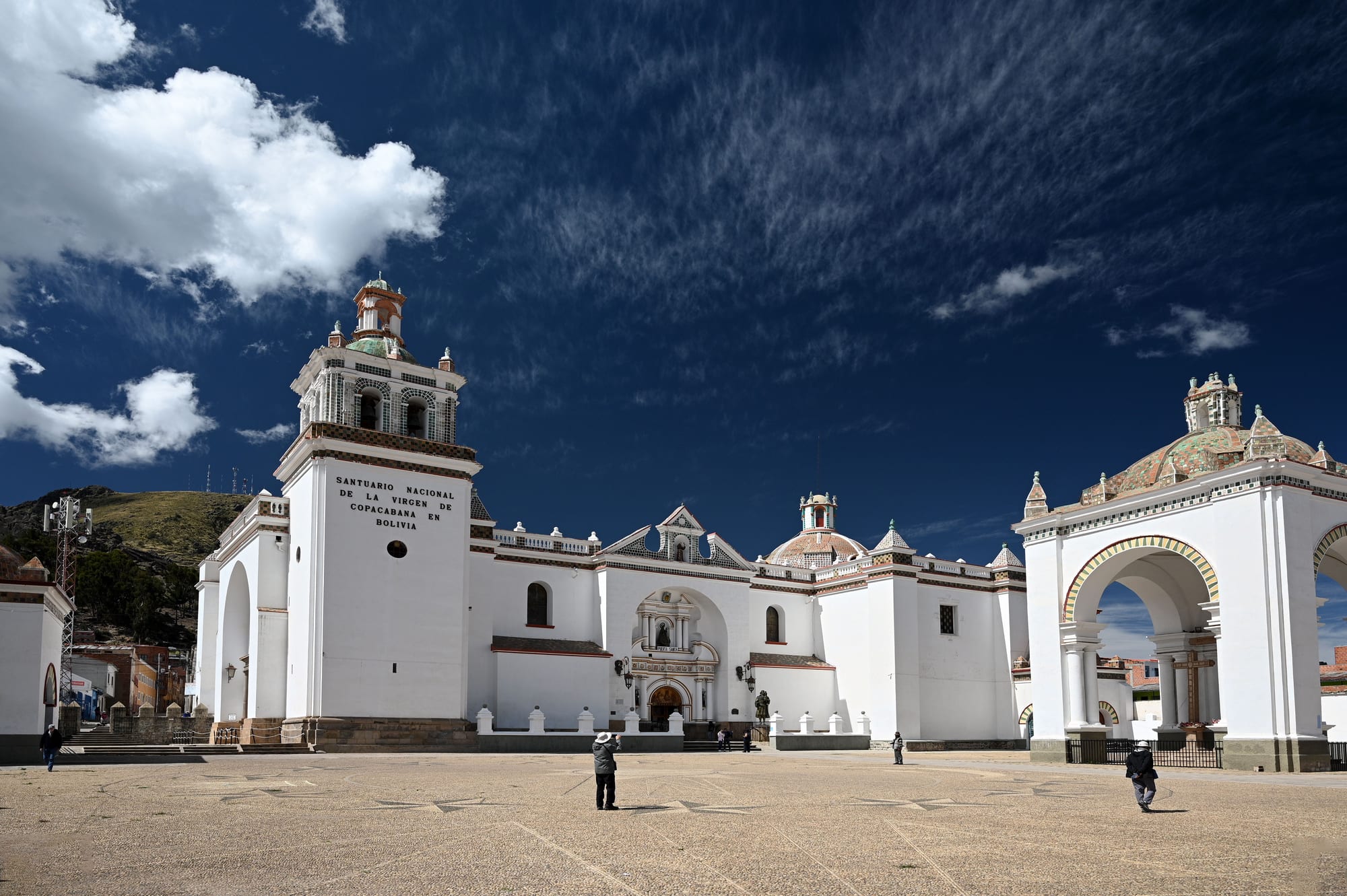 A large white colonial church with a tiled dome and a bell tower bearing the words Santuario Nacional de la Virgen de Copacabana en Bolivia, beside a white arched open-air chapel, under a deep blue sky